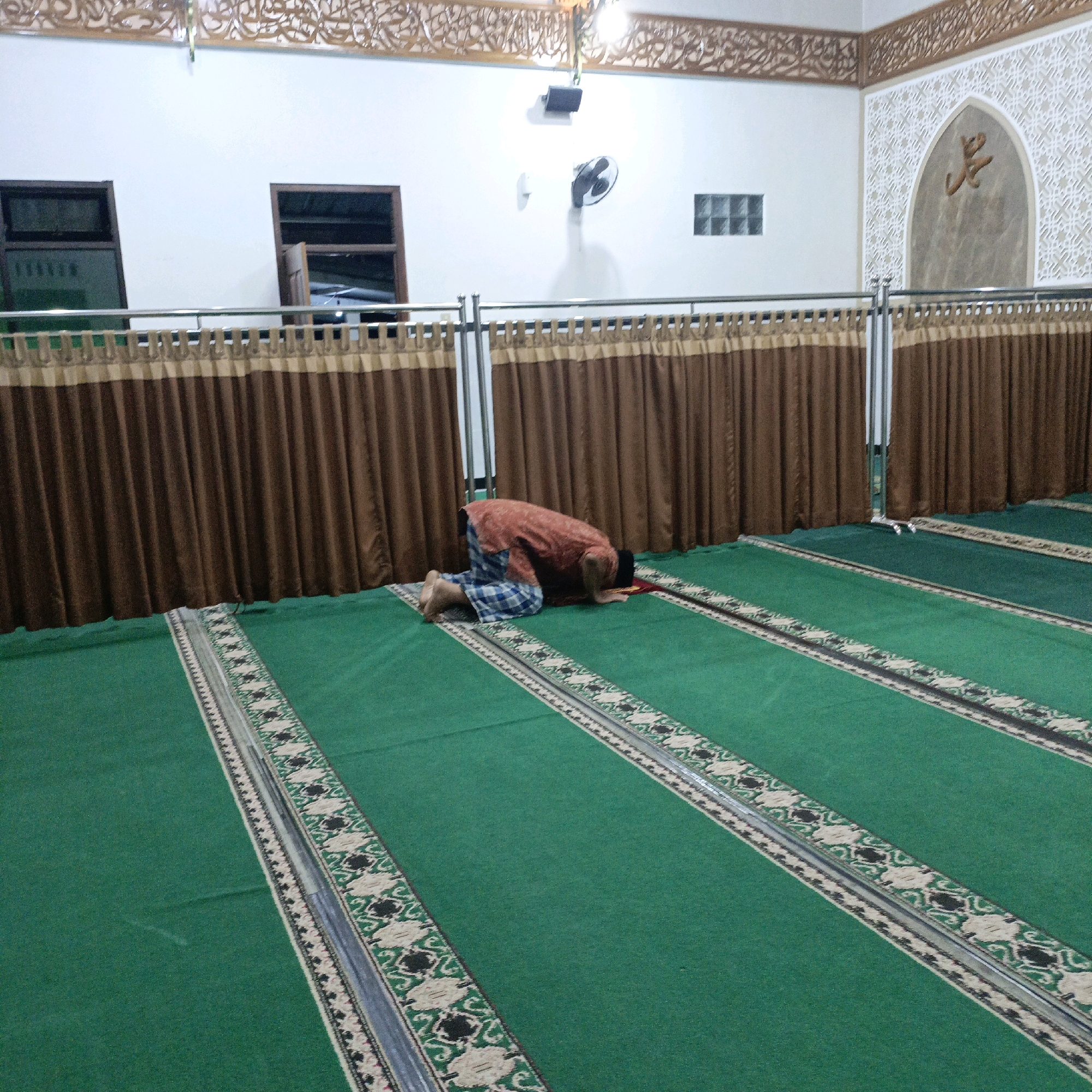 In a mosque in Salatiga, Indonesia, a person wearing a patterned sarong and an orange shirt is in the middle of a prayer. The person is kneeling on a green carpet with a decorative border and bowing their head to the floor. The mosque's interior features brown curtains, white walls, and Arabic calligraphy. A wall fan is mounted on the wall, and there are windows on the left side of the room. The scene appears to be indoors, with artificial lighting.