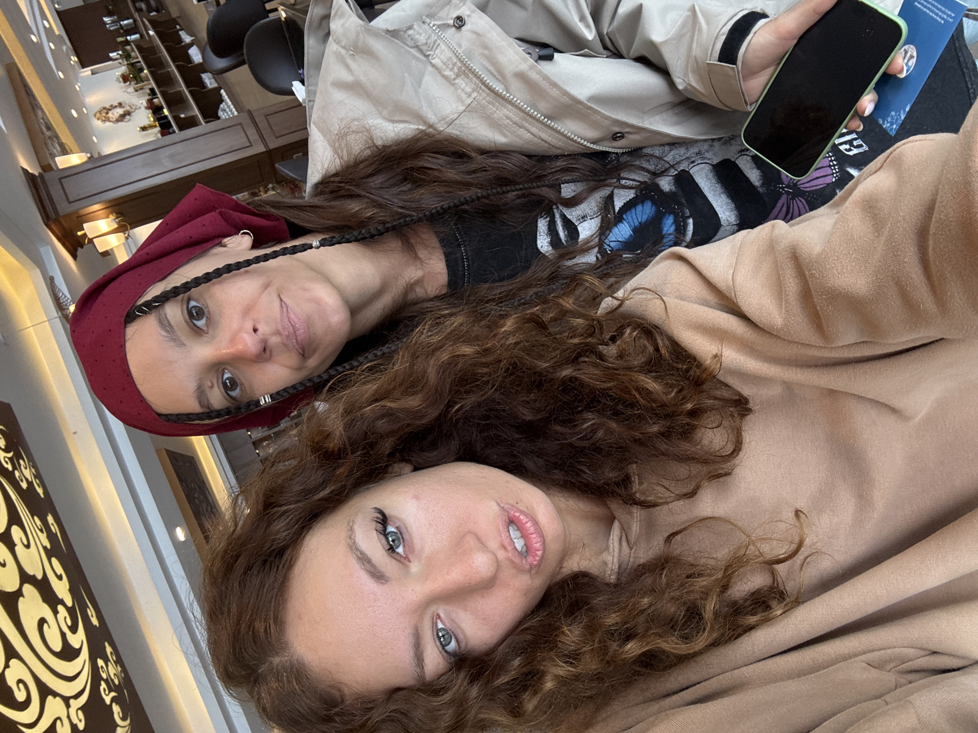 Two women are in Binh Lu, Vietnam, presumably indoors based on the background showing shelving and decor. One woman wears a red head covering and a black shirt with a butterfly graphic under a tan jacket. She has dark hair braided on either side of her face. She is looking into the camera. The other woman is closer to the camera, wearing a tan top, and has curly brown hair. A black smartphone is in the image, held by one of the women. The image appears to be a selfie.