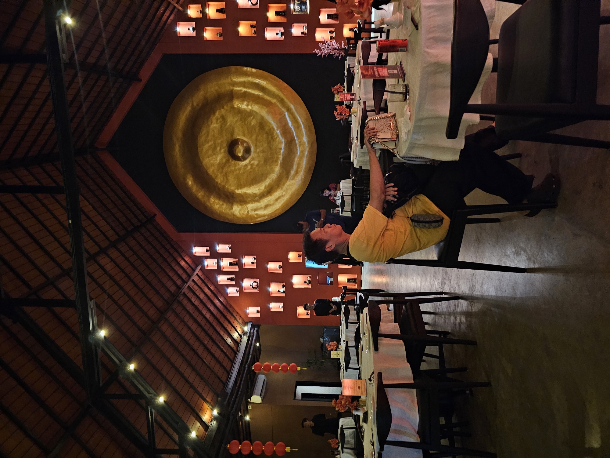 The image shows the interior of a restaurant, likely in Kuta, Indonesia, given the architectural style and decor. The restaurant features a high, sloped, dark wood ceiling adorned with strings of lights and decorative lanterns. A large, round, golden gong is mounted on a black wall. The walls are also decorated with numerous illuminated cubbyholes displaying various objects. A person is seated at a table covered with a white tablecloth, possibly taking a picture with their mobile phone or camera. Other tables and chairs are arranged around the space, suggesting a dining area. Additional red lanterns hang from the ceiling.