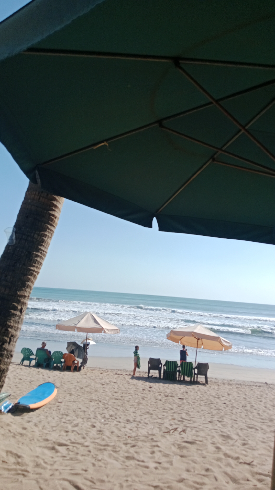 This is a photograph of Kuta Beach, Bali, Indonesia, on a clear day. The foreground is dominated by the texture of wet sand, with a bright blue and orange surfboard lying on it. In the mid-ground, several beach chairs are arranged under two large, tan beach umbrellas. To the left, under one umbrella, a person is seated in a green chair. To the right, under the other umbrella, two individuals are visible, one standing and one appearing to be seated. A child in striped clothing stands near the umbrella on the right. Beyond the beach chairs and umbrellas, the ocean stretches out to the horizon, with gentle waves rolling onto the shore. A palm tree trunk is visible on the far left of the frame. The sky is a clear, pale blue. The image is taken from a low angle, looking up towards the umbrellas and the sky.