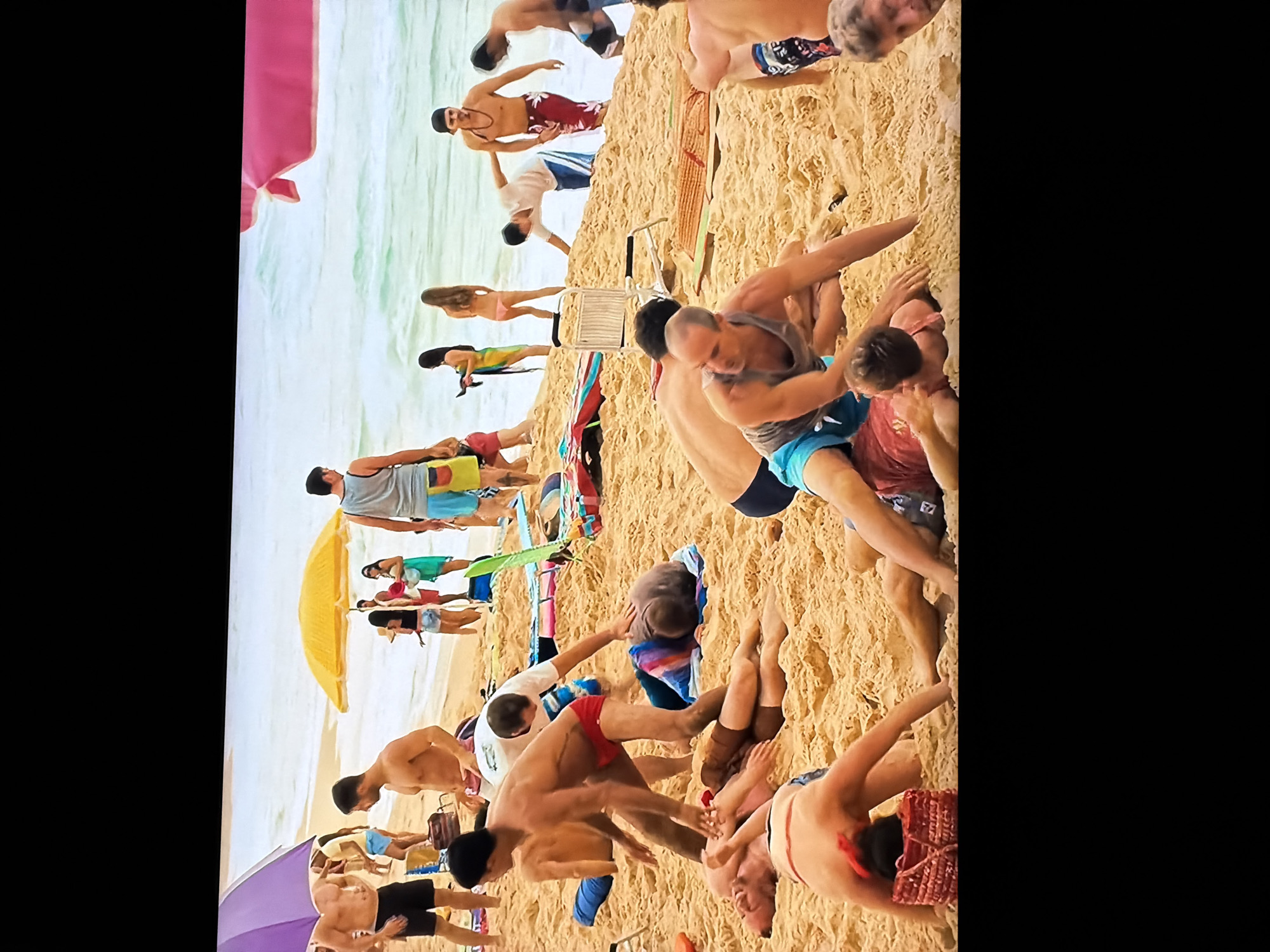 This image depicts a vibrant and densely populated beach scene on a sunny day. The clear, light blue-green ocean stretches across the background, while golden sand fills the foreground and midground.
Numerous individuals, primarily adults and young adults, are present. Many are seen in the water, swimming or wading. On the sand, people are engaged in a variety of activities: some are walking, others are relaxing on towels or beach chairs, and several groups of men are notably clustered together, appearing to be wrestling, playing games, or engaging in other close physical interactions. One person is seen carrying a surfboard near the water's edge.
The beach is equipped with several colorful umbrellas in shades of pink, yellow, and purple, providing shade. Other beach items such as towels, bags, and what appears to be a small portable chair are visible on the sand. Most people are dressed in typical beach attire, including swim trunks and bikinis, with many men shirtless. The bright sunlight indicates it is daytime, likely late morning or afternoon, and the weather appears clear and pleasant. The scene conveys a lively and recreational atmosphere. No visible text is discernible within the image.