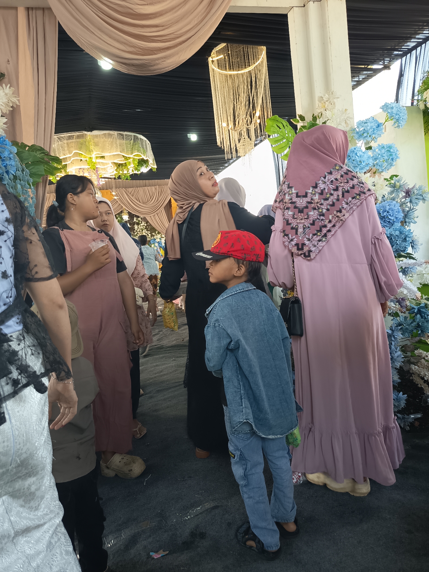 The image depicts an indoor gathering, possibly a celebration or wedding, with an emphasis on fabric decorations and floral arrangements. Several people are visible, including women in modest clothing and a child in a denim jacket and a red cap. The environment appears to be decorated with draped fabric in shades of beige and pink, and large floral arrangements featuring blue and white flowers. A golden chandelier-like decoration hangs from the ceiling. The ground is covered with a dark carpet or tarp. The lighting suggests an artificial indoor setting.