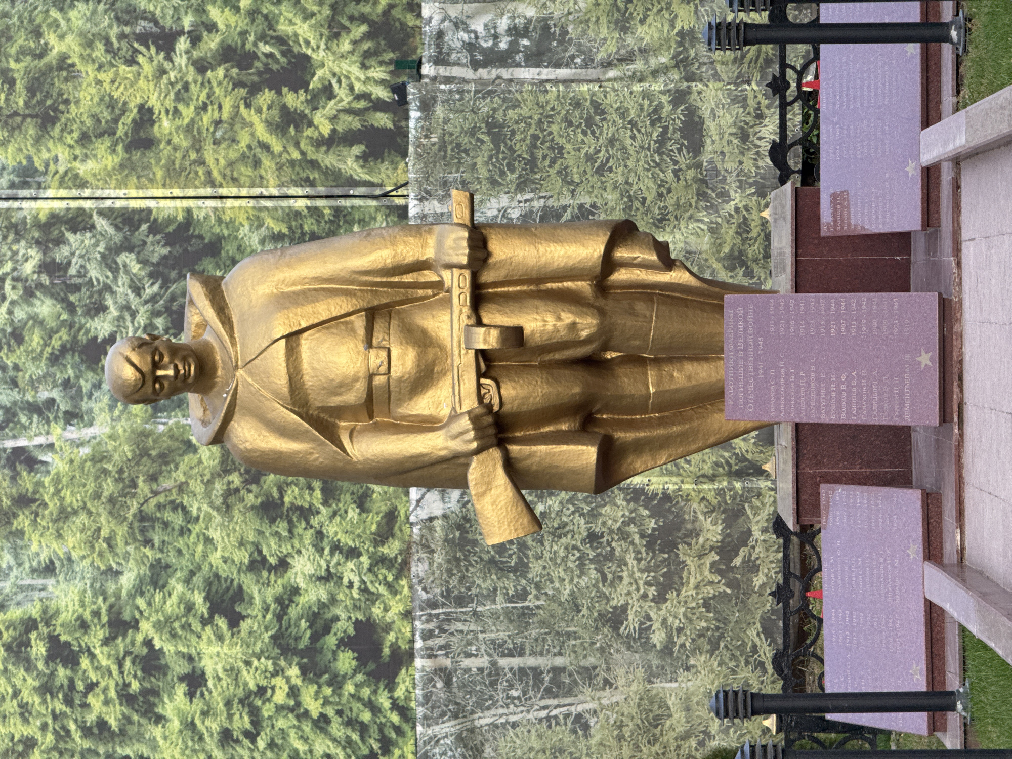 The image shows a golden statue of a soldier holding a rifle, likely a monument or memorial. The statue stands on a stone base with plaques containing inscriptions in Russian. In the background is a screen printed with a dense green forest scene. There are lampposts, and some greenery, around the monument. The location of this monument is Unknown, Unknown.