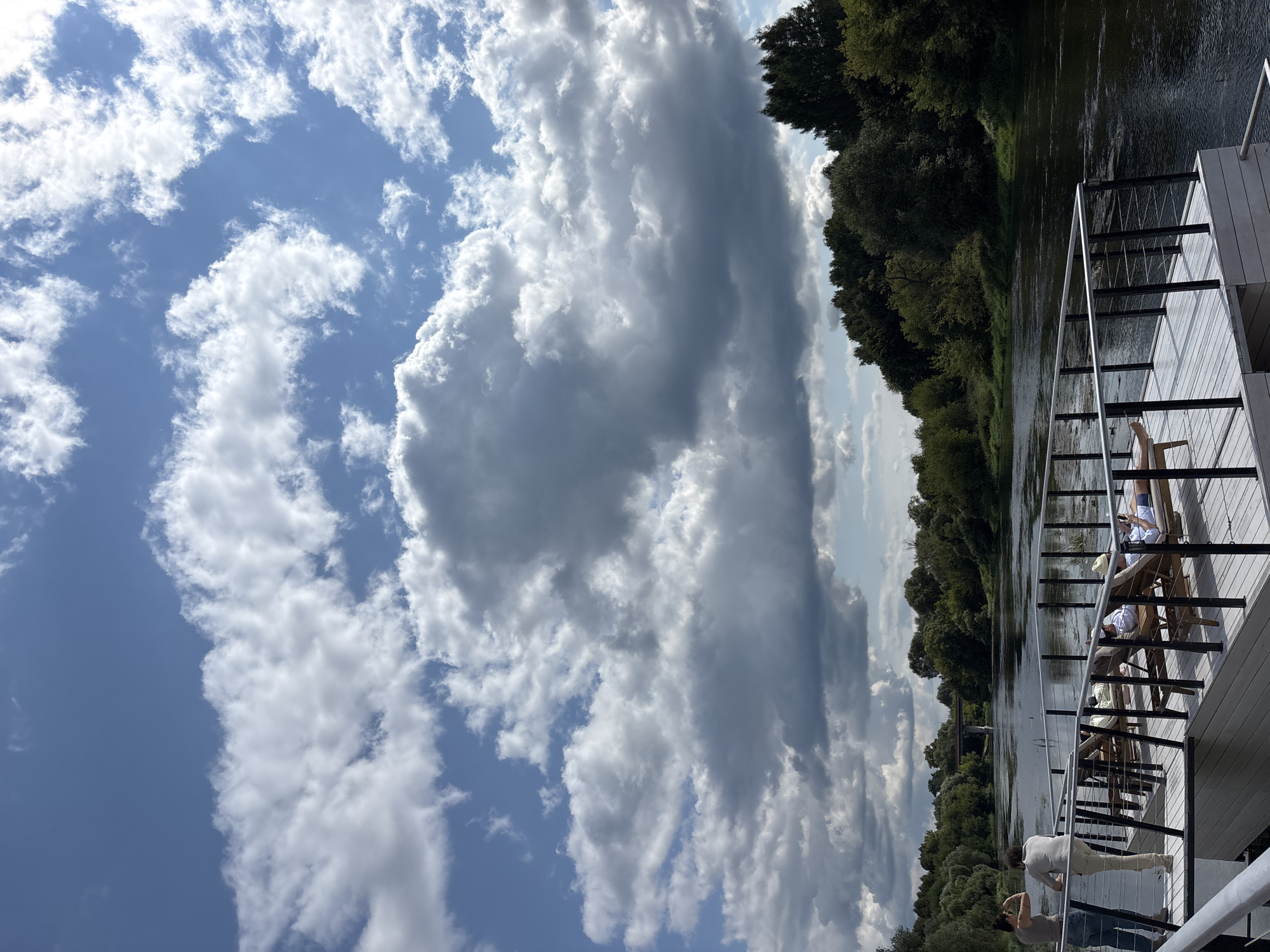 The image depicts a scenic view of the Moskva River in Zvenigorod, Russia, on a partly cloudy day. In the foreground, a white wooden platform extends over the water, equipped with a black metal railing. Several lounge chairs are arranged on the platform, with three people relaxing on them. One individual is lying down, reading a book on a device, while another is reclining with a hat. Two other people are standing at the edge of the platform, looking out at the river.
The river itself is dark and calm, reflecting the sky. Lush green trees and vegetation line both banks of the river, forming a dense forest that stretches into the distance. In the background, a bridge can be seen crossing the river. The sky is a vibrant blue, interspersed with dramatic, voluminous white and gray clouds that create a dynamic atmosphere. The overall scene conveys a sense of tranquility and leisure, set against a beautiful natural landscape.