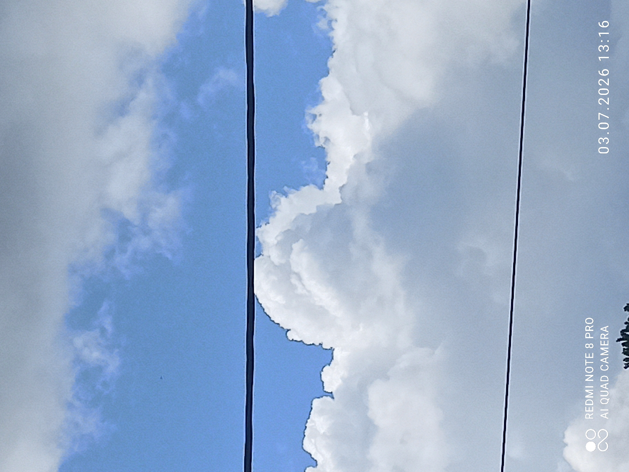 The image is a close-up of a cloudy sky. Two black wires stretch horizontally across the frame, dividing the sky into sections. The top section of the sky is a bright blue, with some wispy white clouds visible. The middle section is dominated by large, puffy white clouds that obscure much of the blue sky. The bottom section is a lighter gray, indicating more distant or less illuminated clouds. In the lower left corner, there is text that reads "REDMI NOTE 8 PRO AI QUAD CAMERA". In the lower right corner, there is text that reads "03.07.2026 13:16". The image does not show any people, buildings, or any indication of the city of Sovetskiy, Russia.