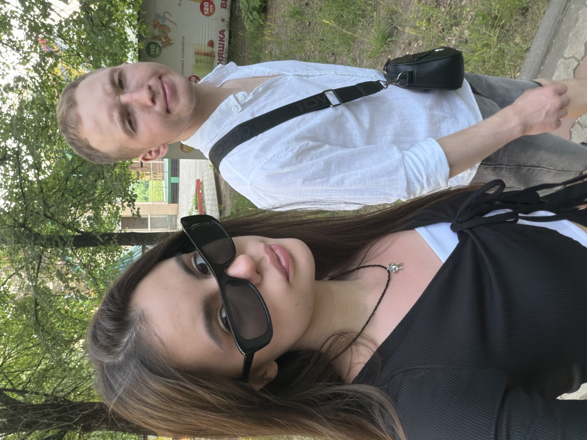 A woman and a man are posing for a selfie outdoors. The woman is in the foreground wearing dark sunglasses, a black top, and a necklace with a pendant. Her long, brown hair is parted to the side. The man is positioned behind her and to the side, wearing a white button-down shirt, grey pants, and a black crossbody bag with a black strap.
The background shows a green, leafy environment, suggesting a park or a street lined with trees. Behind the man are some structures, including what appears to be a low building and a promotional sign for a drink or dessert called "НЯШКА". The sign also contains information about "120 РУБ".
The city is Chaikovsky, Russia.