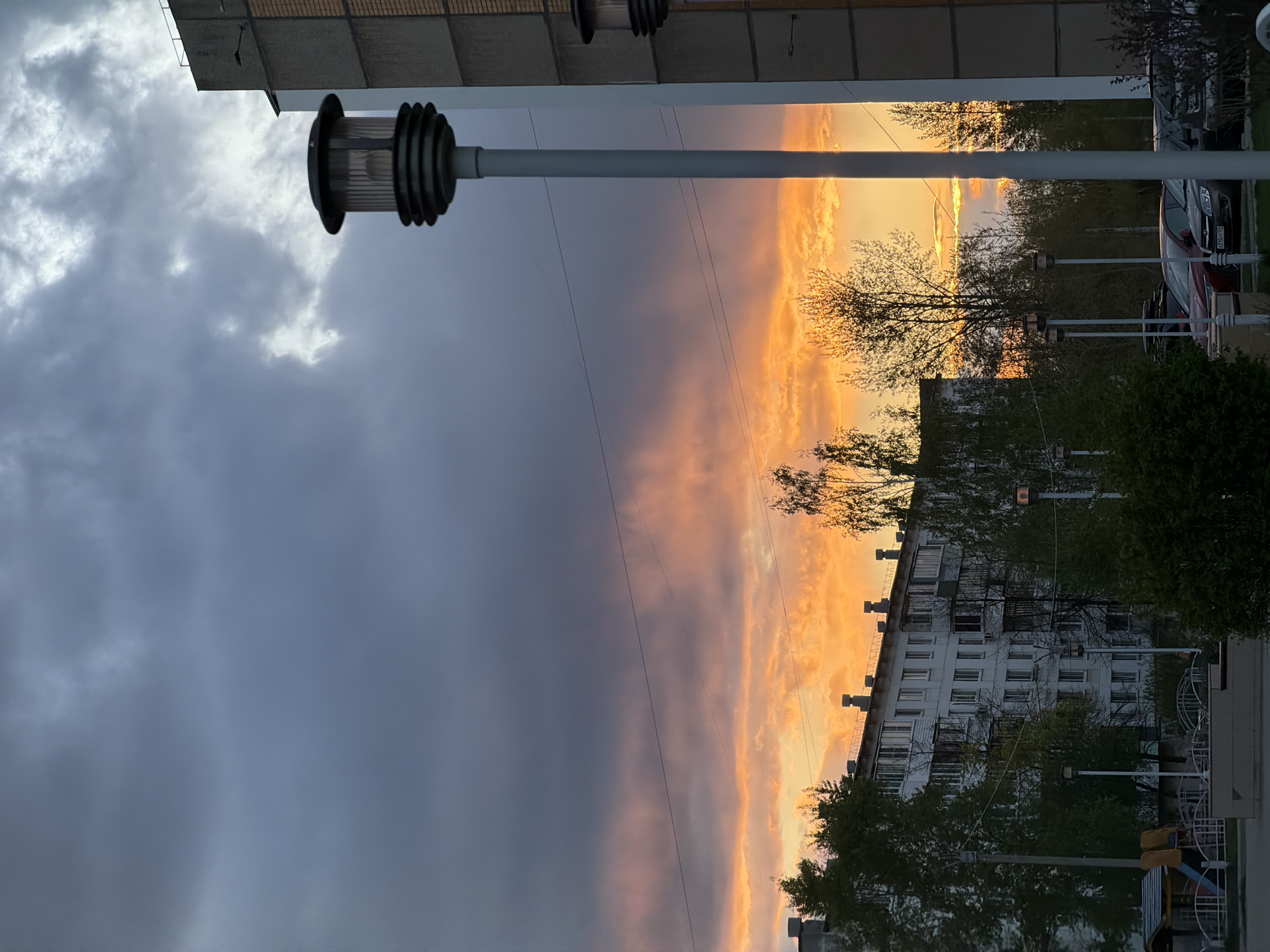 The image depicts a sunset over a cityscape in Chaikovsky, Russia. A gray sky occupies the left portion of the frame, contrasted by a band of orange and yellow clouds where the sun is setting in the distance. Buildings, trees, and streetlights are silhouetted against the colorful sky. Cars are visible on the right. The image is taken from a low angle, looking upwards.