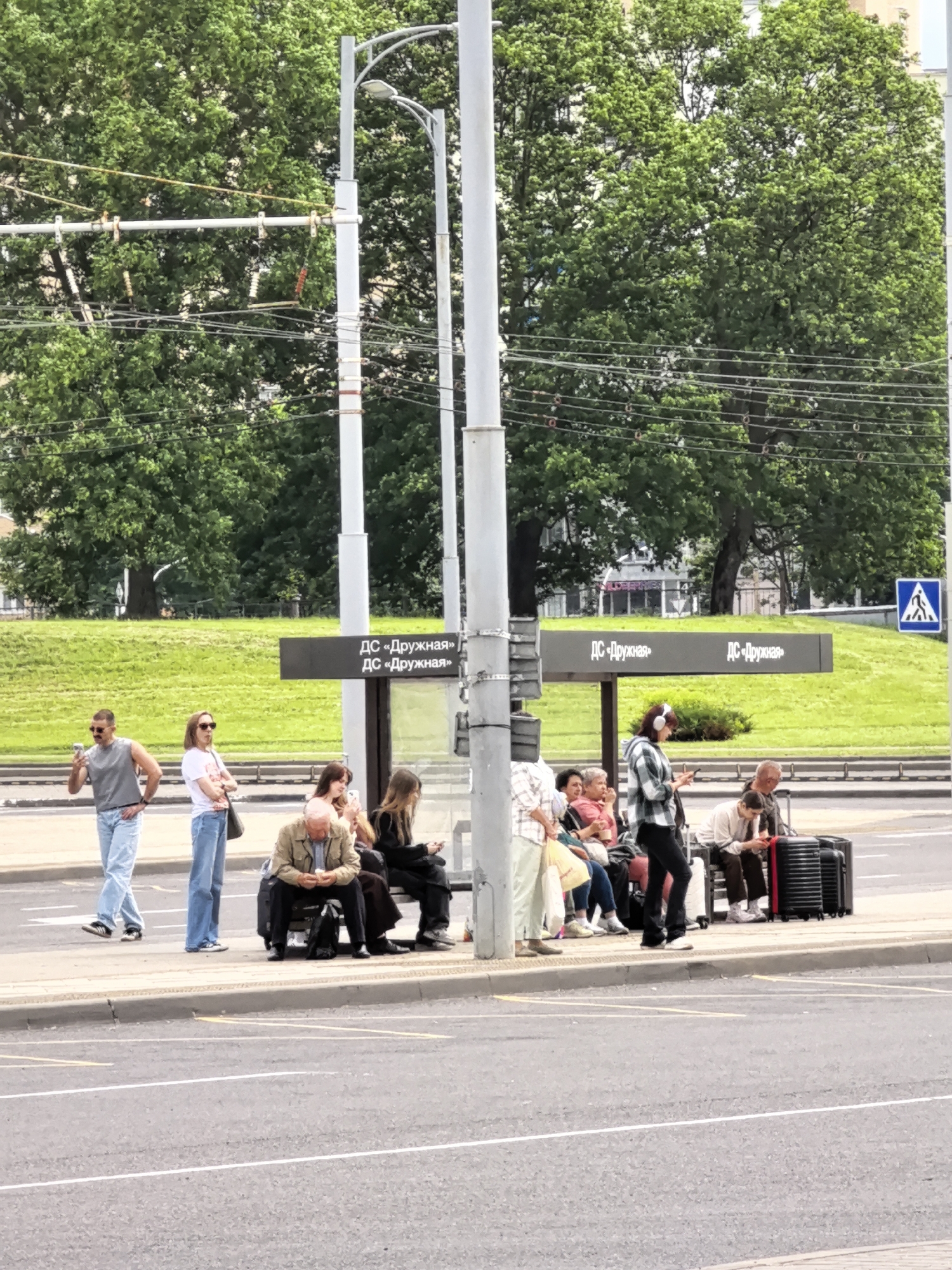 This media file depicts a daytime scene at a bus stop, possibly in an urban or suburban setting, given the presence of a bus shelter and overhead wires. A diverse group of people are waiting at the bus stop, with some seated on a bench and others standing. They appear to be casually dressed, suggesting a warm day. Many are engaged with their phones, while others are looking around or conversing. One person is wearing headphones. Luggage is visible near some of the individuals, indicating they may be traveling. The bus shelter has dark signage with Cyrillic text that reads "ДС «Дружная»" repeated twice on each side. In the background, there are trees with lush green foliage and a grassy embankment. The overall atmosphere seems relaxed and ordinary, typical of a public transportation waiting area. The presence of a pedestrian crossing sign in the upper right corner further suggests a public road or intersection. The weather appears to be overcast or partially cloudy, as the lighting is diffused.