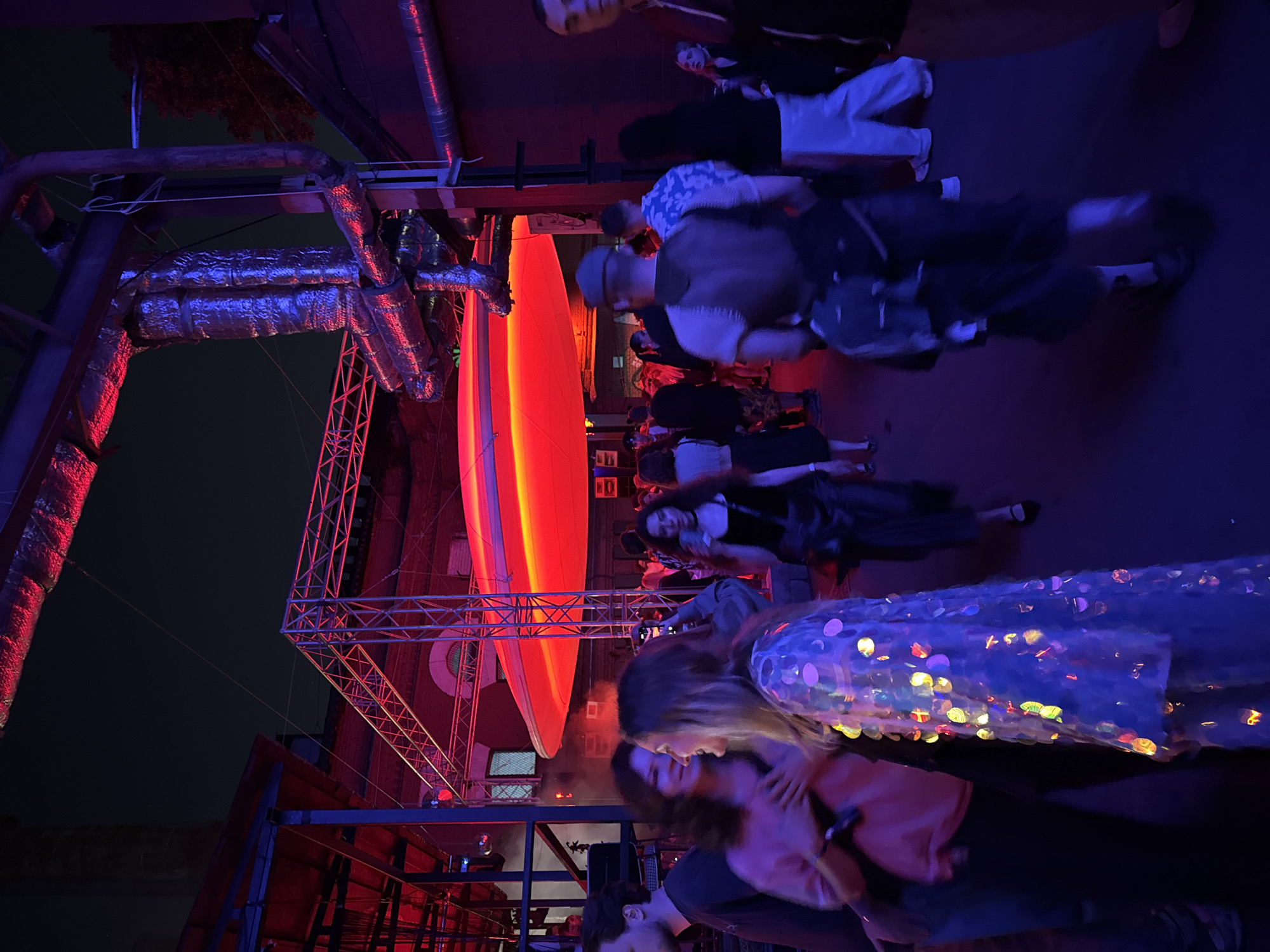 The image depicts a crowded outdoor or semi-outdoor event space at night, identified as the Mutabor club in Moscow, Russia, due to the distinctive large, glowing red oval light fixture suspended overhead, which is a signature architectural element of the venue located on Yauzskaya Street.
The scene is illuminated predominantly by artificial red and blue light, creating a dynamic, low-light environment. Overhead, large, insulated industrial pipes and metal truss structures are visible, consistent with a repurposed industrial aesthetic. In the mid-ground, a prominent, elongated, horizontally oriented red light fixture, resembling a suspended elliptical disc, emits a strong red glow, illuminating the area beneath it.
Numerous individuals are present, socializing and moving through the space. In the immediate mid-foreground, a woman with long blonde hair, wearing a shimmering, sequined garment that reflects various colors (blue, yellow, pink, green) from the ambient lighting, is smiling and looking towards her right. To her left, another woman with dark, curly hair, wearing a pink top, is smiling and facing the blonde woman, with her right hand lightly placed near the blonde woman's shoulder. Further to the left, a man's partial profile is visible.
In the mid-ground to the right, a man wearing a cap and a dark vest is seen from the back, walking forward. The background shows a dense crowd of people, many of whom are blurred due to motion or shallow depth of field, suggesting a lively event. Architectural details, including arched windows and brick-like walls, are dimly visible in the background, further confirming the industrial setting. The overall atmosphere is indicative of a nocturnal social gathering or party.
