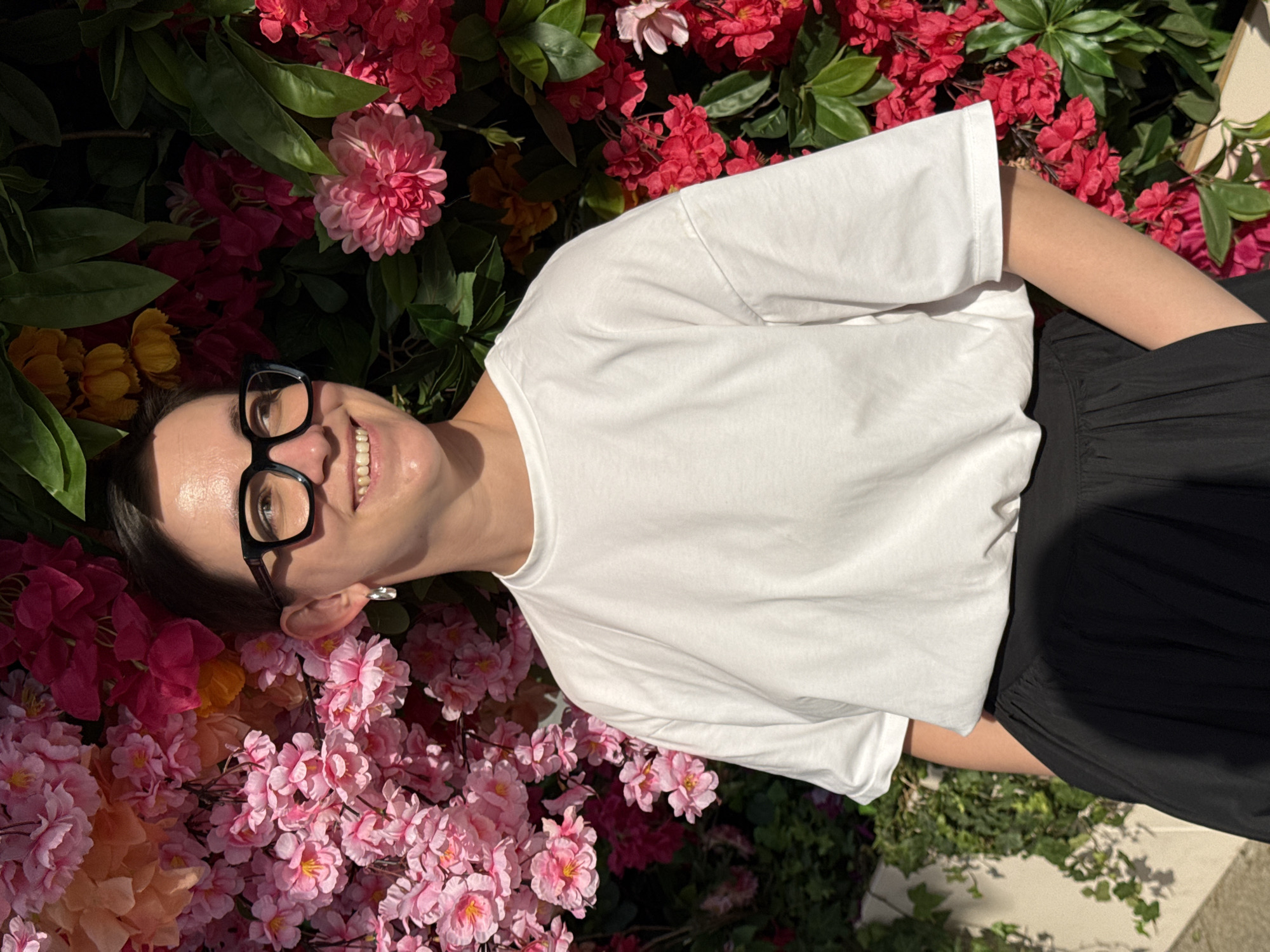 A person wearing glasses and a white t-shirt is smiling while standing in front of a floral wall with red, pink, and orange flowers. The person is also wearing black pants. The location is difficult to identify, but the image does not clearly show characteristics that would help determine if it is in Moscow, Russia.