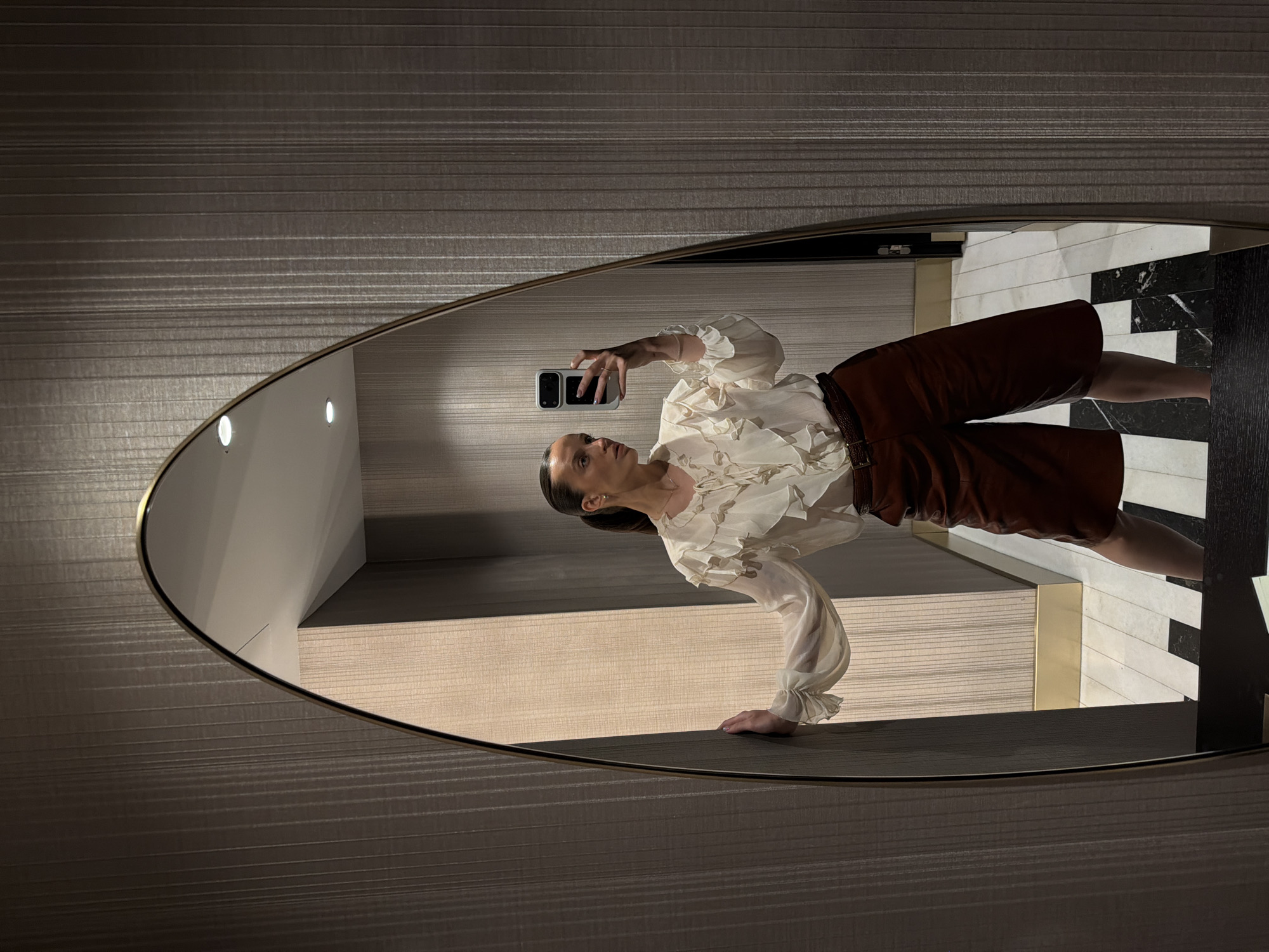 A woman takes a selfie in a mirror. She is wearing a white ruffled blouse and brown shorts. The mirror is oval and framed in gold. The background is a wall with a vertical ribbed texture. The floor is tiled with black and white geometric patterns. The scene appears to be indoors, possibly in a hotel room or a fitting room. The location is not Budva, Montenegro.