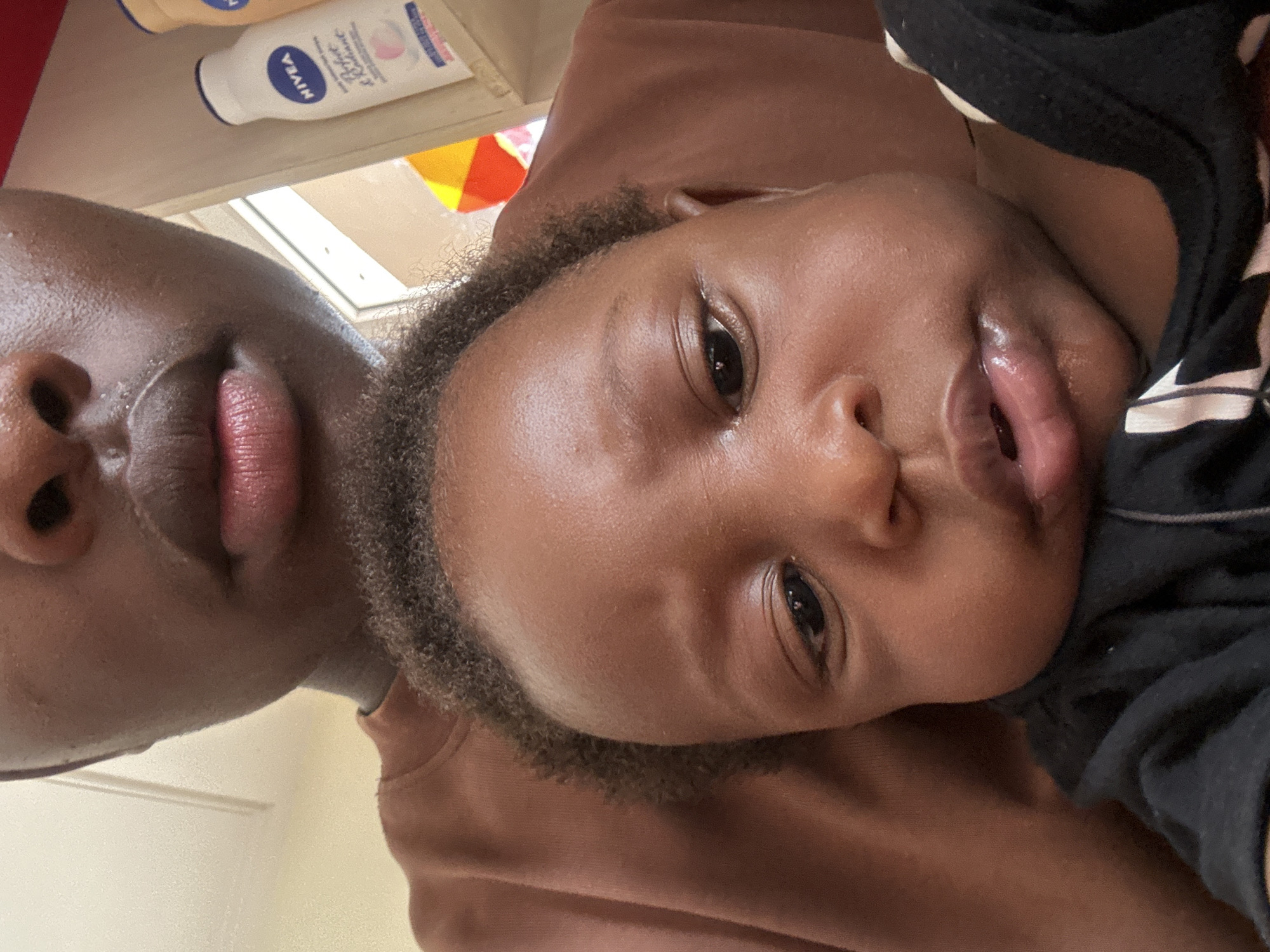 This close-up, rotated image features two individuals with dark skin tones: an adult (partially visible on the left) and a young child (more fully visible on the right), likely engaged in a selfie or close-up interaction. Both subjects are looking towards the camera.
The adult's face is cropped, showing the nose and lips, which have a reddish hue. The child's face is prominent, featuring wide, dark eyes, a slightly open mouth, and short, dark, curly hair. The child appears to be wearing a black garment, possibly with white stripes, visible on the right side.
In the background, positioned above the subjects, there is a shelf or surface holding two bottles. One is a white Nivea lotion bottle, clearly displaying the text "NIVEA" and "Soft & Créme." A second, yellow or orange bottle is partially visible beside it. The bright background suggests an indoor setting with ample natural light, possibly near a window. The overall mood is intimate and gentle, capturing a moment of closeness between the adult and child. The scene likely takes place in Jos, Nigeria.