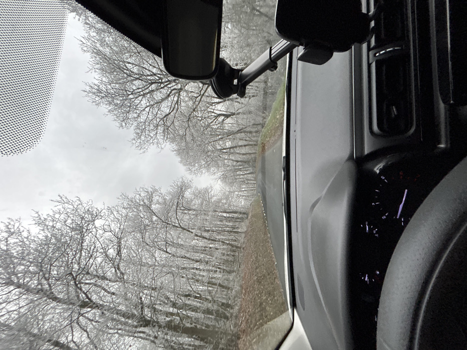 The image depicts a driver's perspective from within a vehicle on a rural road in Saint-Romain-en-Gal, France, during winter conditions. The foreground reveals the dark dashboard, a partial view of the steering wheel, and an illuminated instrument cluster on the left. A black phone mount with an adjustable arm is suction-cupped to the right side of the windshield, below the rearview mirror, which is also visible. The upper windshield area features a black dot matrix pattern.
The road ahead is asphalt, dark gray, and appears to curve gently to the left while ascending. Small, white markers line the left edge of the roadway. Both sides of the road are bordered by dense deciduous trees, all of which are bare of leaves. Their branches and twigs are heavily coated in a visible layer of white frost or ice, creating a stark, monochromatic landscape. The ground alongside the road is covered with brown leaf litter and some sparse green vegetation. The sky is uniformly overcast and light gray, indicating a dull, cold day. No individuals or other vehicles are visible in the scene.