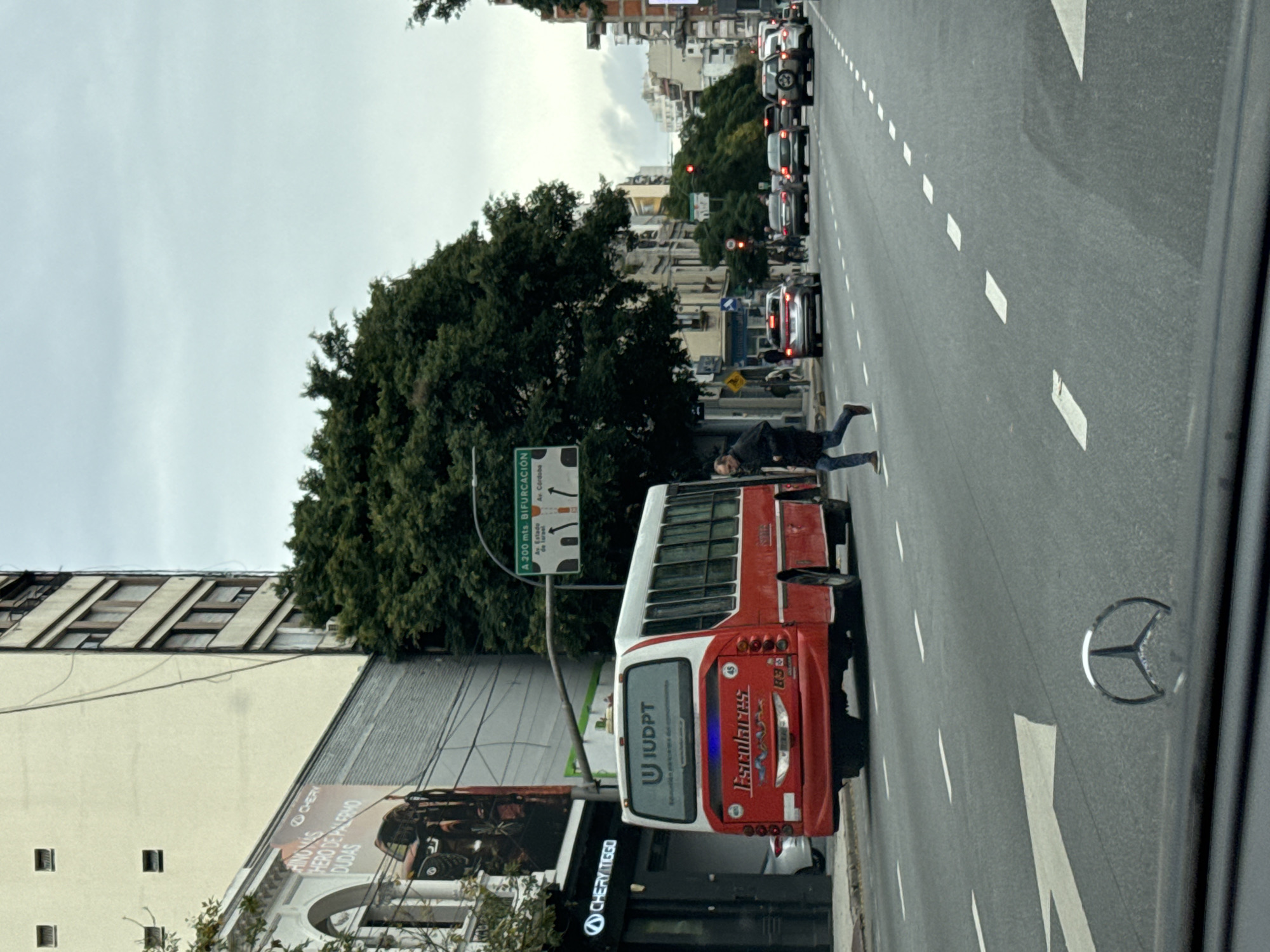 The image shows a street scene in Buenos Aires, Argentina. A red and white school bus is stopped on the right-hand side of the road, occupying a traffic lane. A person is lying on the road near the bus. There are several cars lined up behind the bus, seemingly stopped in traffic. The street is marked with white lane dividers and directional arrows. A road sign indicates "A 200 mts. Bifurcación" followed by "Av. Córdoba." A building on the left has an advertisement for "Chery Tiggo" featuring an off-road vehicle. The advertisement also says "Hero de Palermo" and "Rino Mas Duras." The rear of the bus features "Escolares" and the acronym "IUDPT". A Mercedes-Benz logo is visible in the bottom right corner. The sky is overcast.