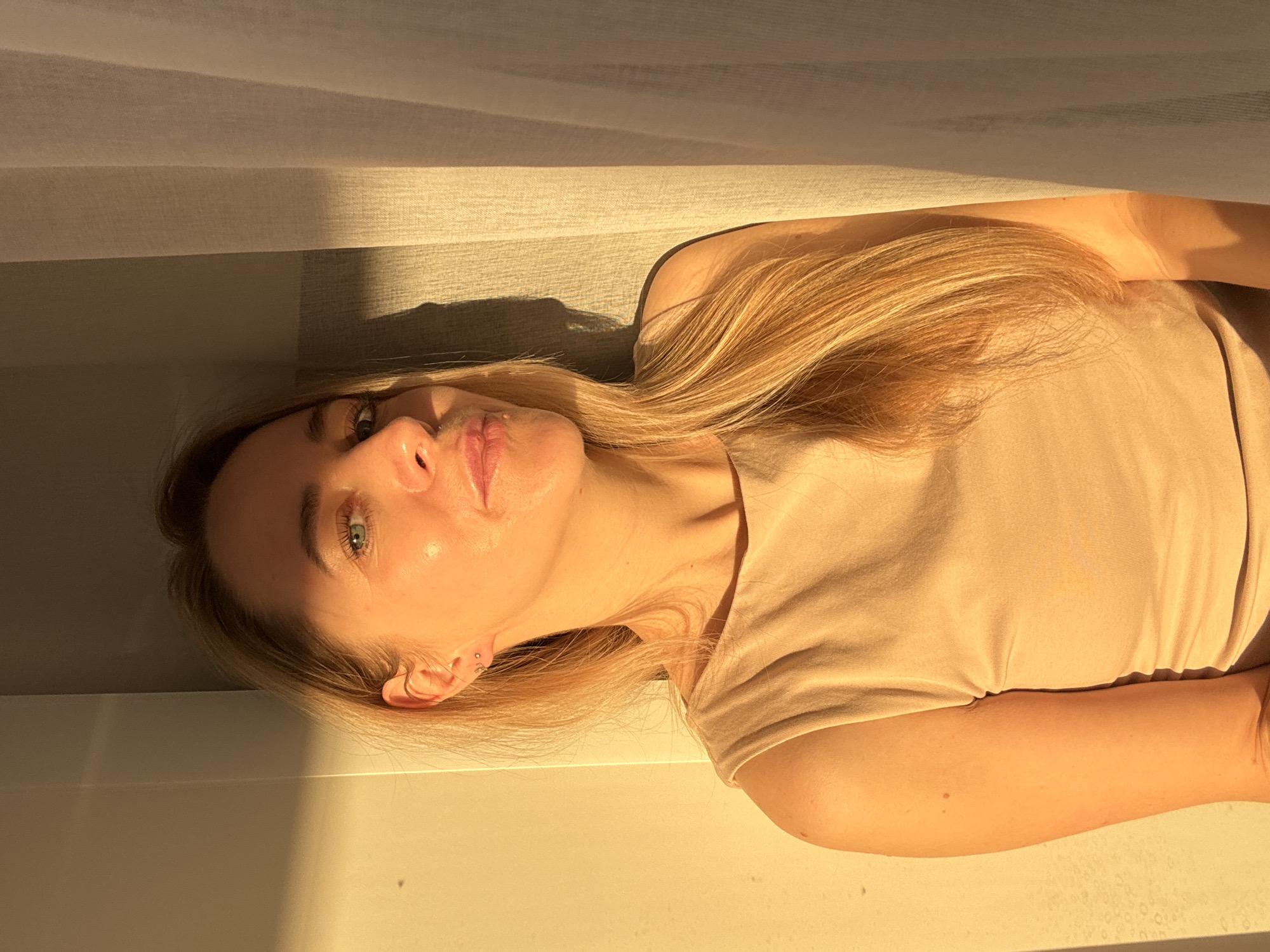 A woman with long, blonde hair is lying down in a room with sunlight streaming through a window covered by blinds. She is wearing a light brown shirt. Her face is visible, and she appears to be looking up. The scene is brightly lit by the sun. There is no clear indication of the location being Chaikovsky, Russia.
