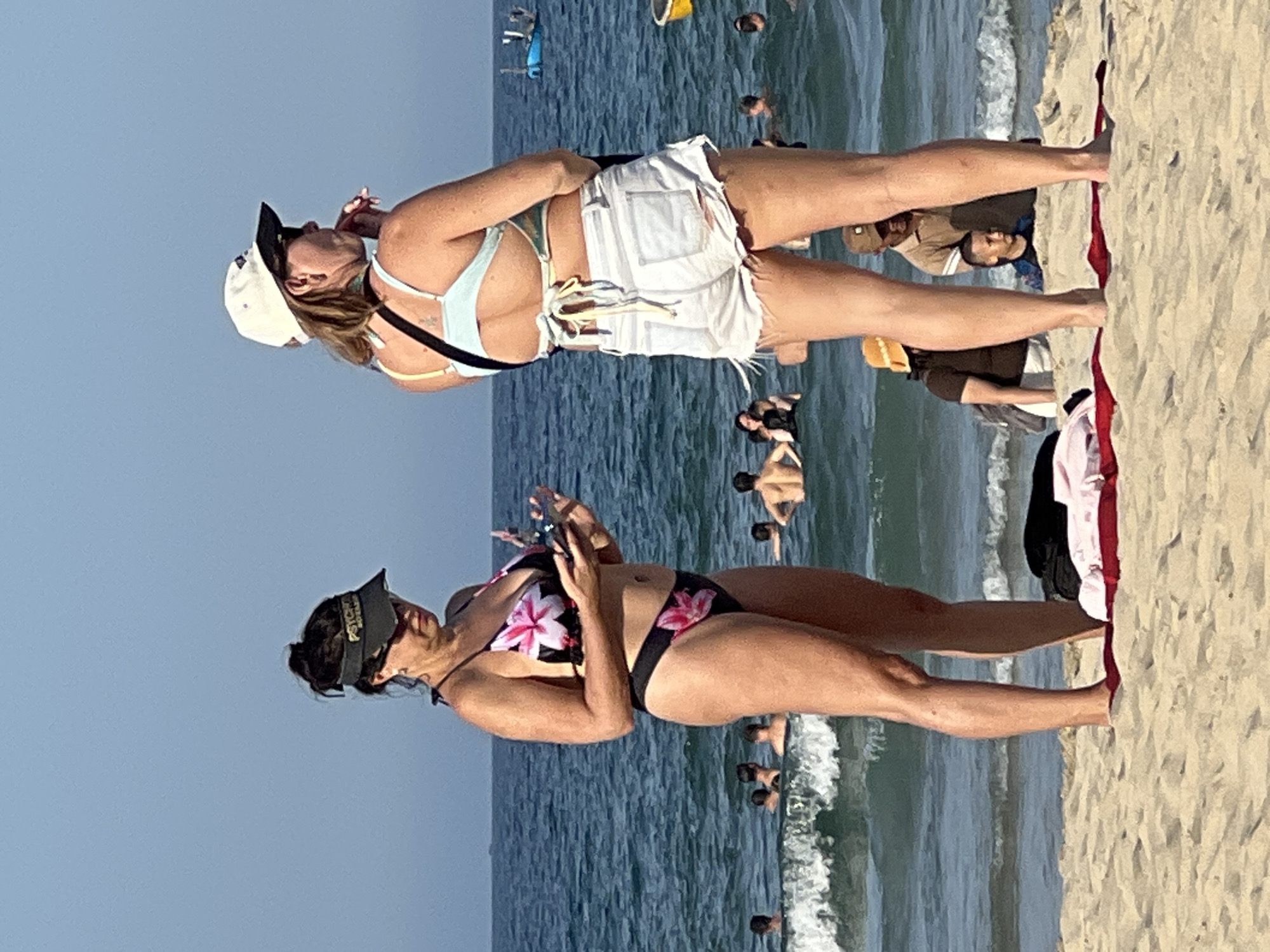 Two women are standing on a sandy beach in Da Nang, Vietnam. The woman on the left is wearing a black and pink floral bikini and a black visor. She is looking down at her phone. The woman on the right is wearing a light blue bikini top and white denim shorts. She is wearing a white baseball cap and appears to be looking out at the ocean, possibly chewing on something. In the background, several people are swimming and wading in the ocean. The water is blue and there are small waves breaking on the shore. The sky is clear and blue.