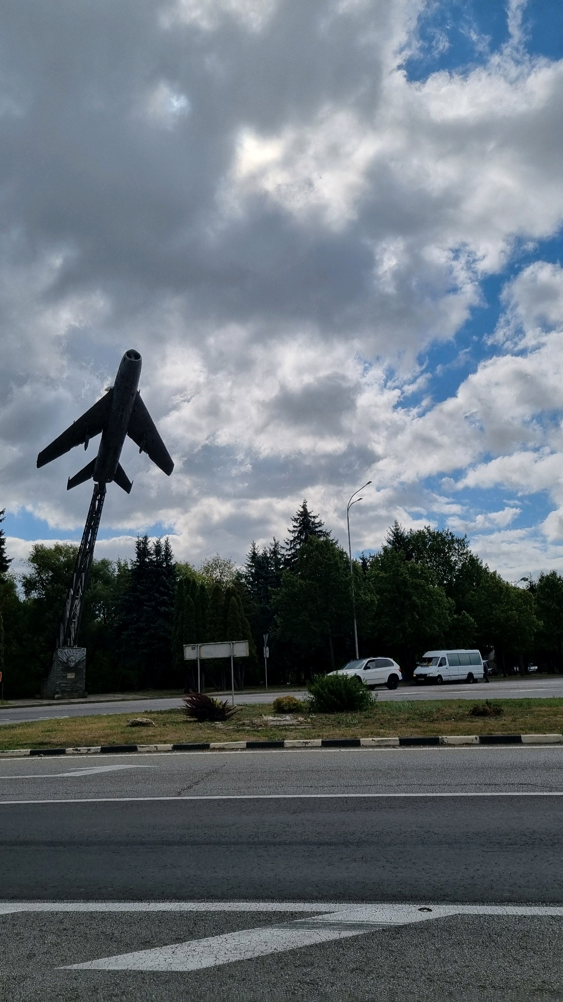 In Dubno, Ukraine, an outdoor scene captures an airplane monument against a backdrop of a cloudy sky. The monument, a dark, silhouetted jet, is mounted on a tall, slender metal pylon, tilted upwards as if in flight. The pylon is supported by a stone base. To the right of the monument, a row of green trees forms a dense line, with a street light visible above them. Beyond the trees, a white car and a white van are parked on the side of a road. The road in the foreground is asphalt, with white lane markings. The sky is filled with a mix of dark grey and bright white clouds, with patches of blue sky visible through the openings. The overall atmosphere is overcast, suggesting a potentially rainy or stormy day.
