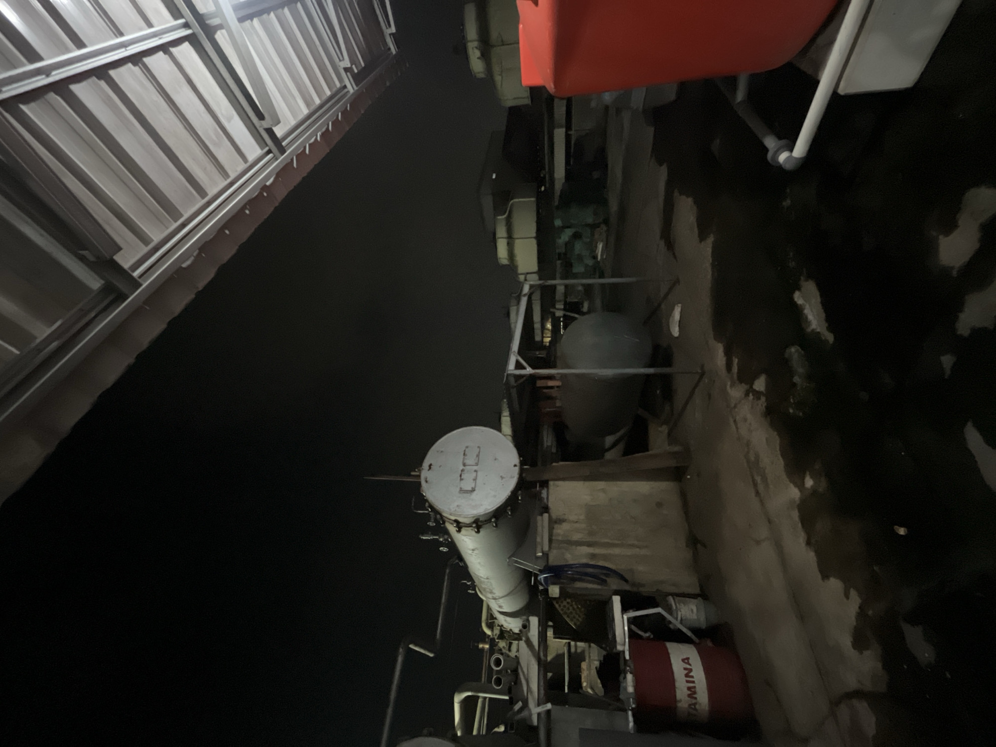 The image depicts an outdoor industrial scene at night, likely on a rooftop. In the foreground, a red barrel with the word "AMINA" is visible, along with a large red plastic water tank on the right side. A large, cylindrical metal tank, possibly a boiler or pressure vessel, is prominently featured in the mid-ground, supported by wooden structures. Behind it, a smaller, gray cylindrical tank is visible. Further in the background, several large industrial cooling towers are present. The ground appears wet and dark, suggesting it might have rained or there's standing water. The scene is illuminated by artificial lighting. The image does not show any people. The overall impression is of an operational or storage area within an industrial facility in Pekalongan, Indonesia.