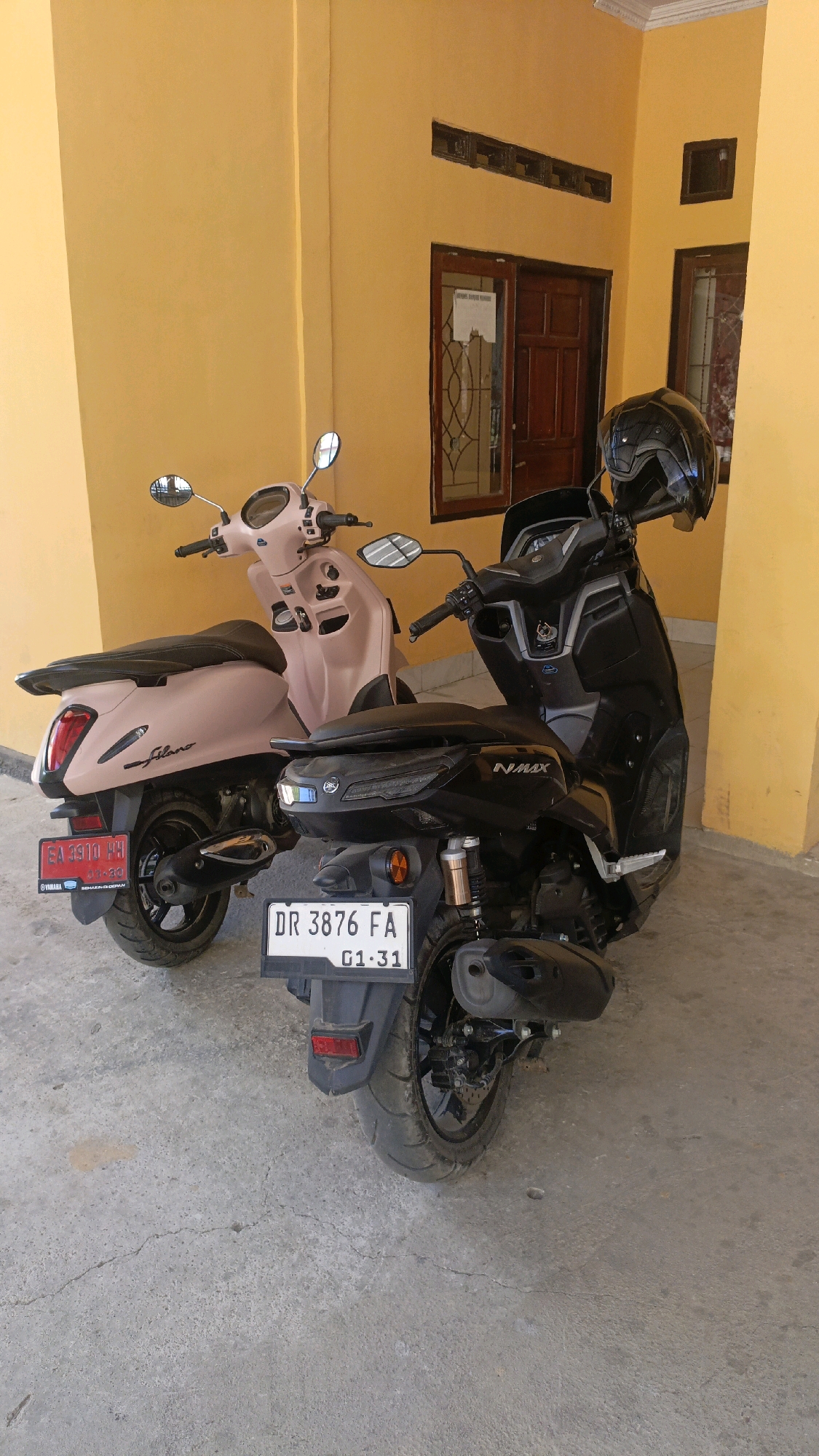 Two scooters are parked on a concrete surface in front of a building with yellow walls and brown wooden doors and windows. The scooter on the left is a light pink scooter with the brand name "filano" visible on its side. It has a license plate that reads "EA 3310 HH" and has a "YAMAHA" logo. The scooter on the right is black and has the brand name "NMAX" visible on its rear. Its license plate reads "DR 3876 FA" and "G1 31". A black helmet rests on the handlebars of the black scooter. The scene appears to be an outdoor area adjacent to a building.