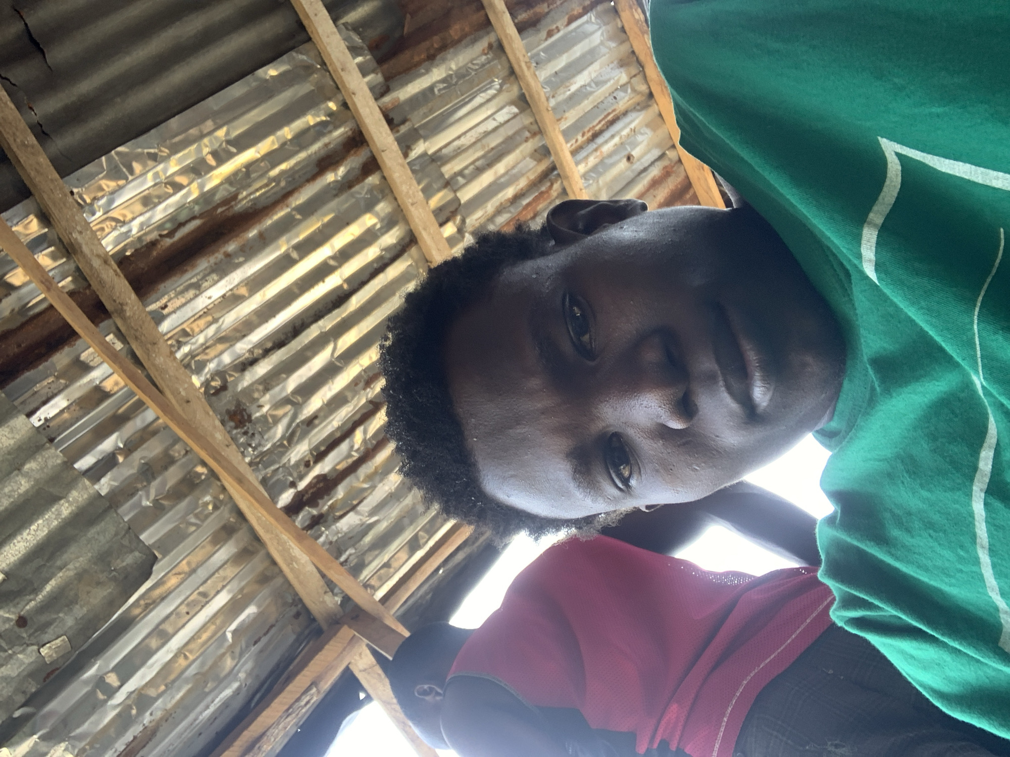 The image shows a selfie taken from a low angle, looking up towards a corrugated metal roof supported by wooden beams. In the foreground, a young person wearing a green t-shirt with white stripes is looking at the camera. Their face is partially visible, with their right eye and forehead in focus. Behind them, another person wearing a pink tank top and possibly dark shorts is visible from the shoulders up. The environment appears to be outdoors, likely under a makeshift shelter. The lighting suggests it is daytime. The overall impression is a casual, candid moment captured in an informal setting within the city of Lekki, Nigeria.