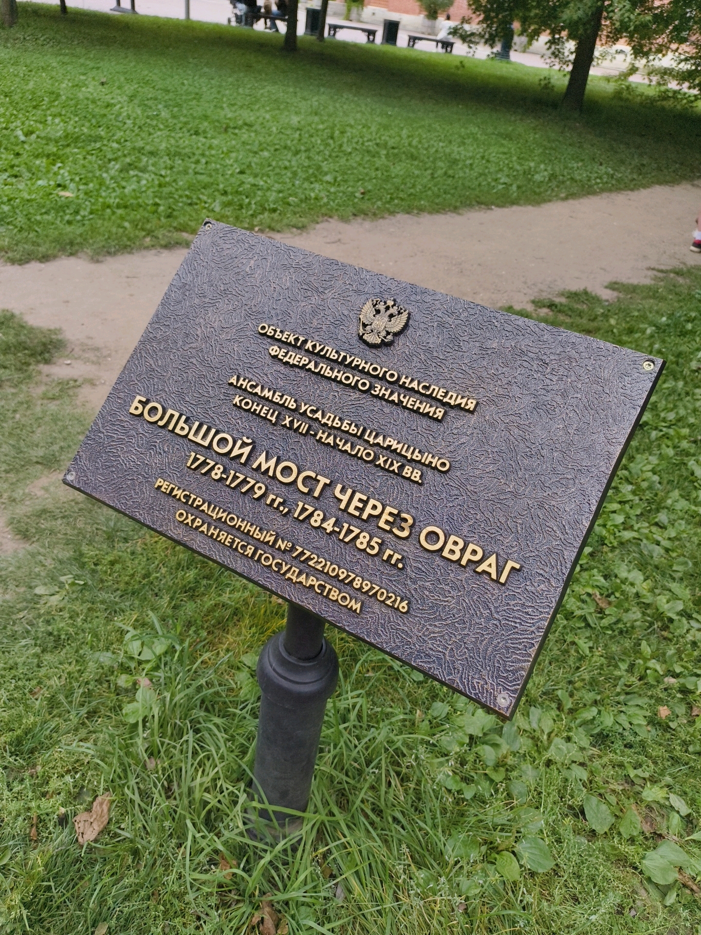 A bronze plaque with embossed Russian text and the Russian coat of arms is mounted on a dark metal pole in a grassy area. The text on the plaque indicates that it is a cultural heritage site of federal significance, an ensemble of the Tsaritsyno estate, late 17th - early 19th centuries. It also states that "Bolshoy Most Cherez Ovrag" (Big Bridge Across Ravine) was built between 1778-1779 and 1784-1785, with a registration number of 772210978970216, and is protected by the state. The background shows a park-like setting with a dirt path, green grass, and trees. A few blurred figures are visible in the distance.