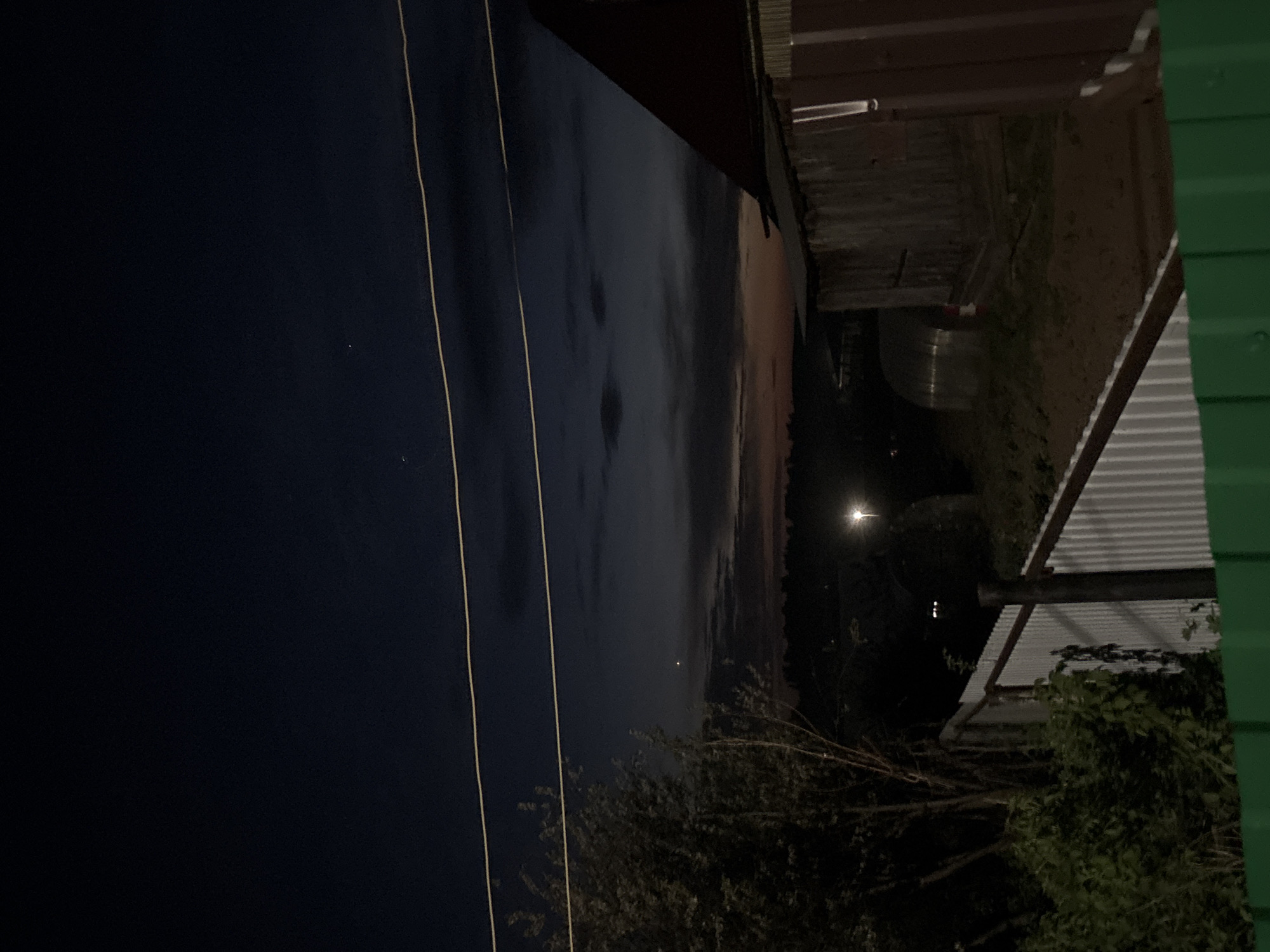 Here is a description of the image:
The image shows a nighttime scene in Фоки, Russia. In the distance, there is a faint sunset on the horizon. In the foreground, there are power lines and trees. To the right, there are buildings, including one with a white corrugated metal roof and another with a green wall. A streetlight illuminates part of the scene.