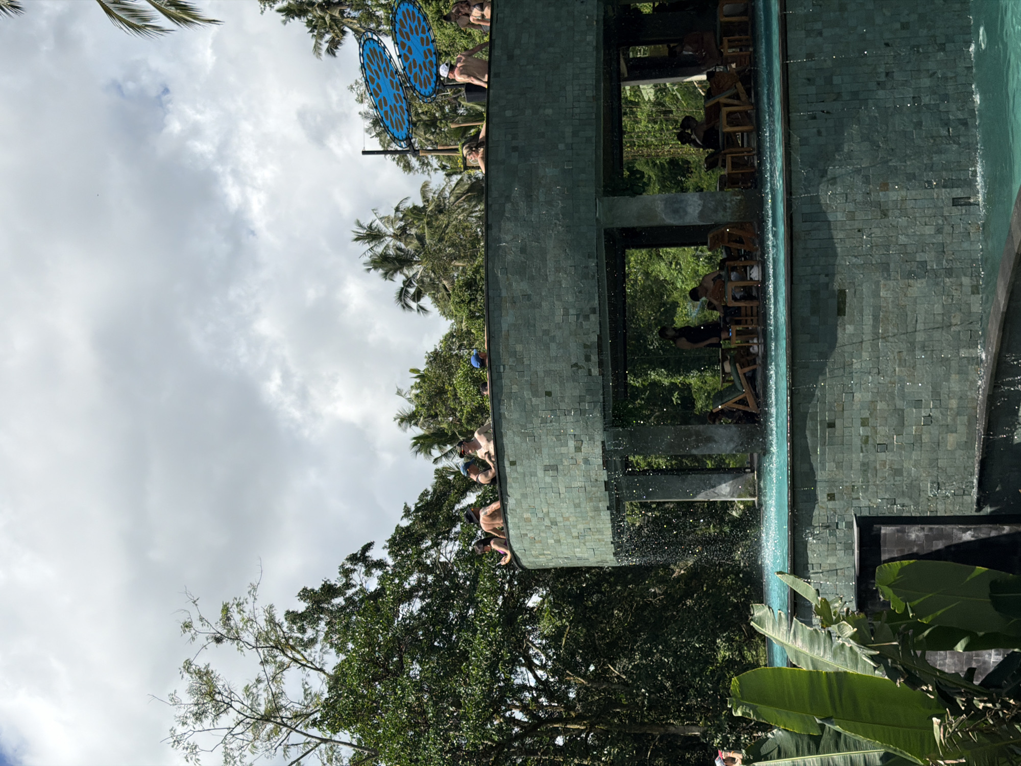 The image shows a multi-level structure with a pool in Tegal Suci, Indonesia. The structure has a green tiled exterior and overlooks a lush, green landscape with trees and vegetation. People are present on the upper levels of the structure, some appearing to be at a bar area with seating. The pool extends along the side of the structure. Blue decorative circular objects are visible in the background. The sky is overcast with clouds.