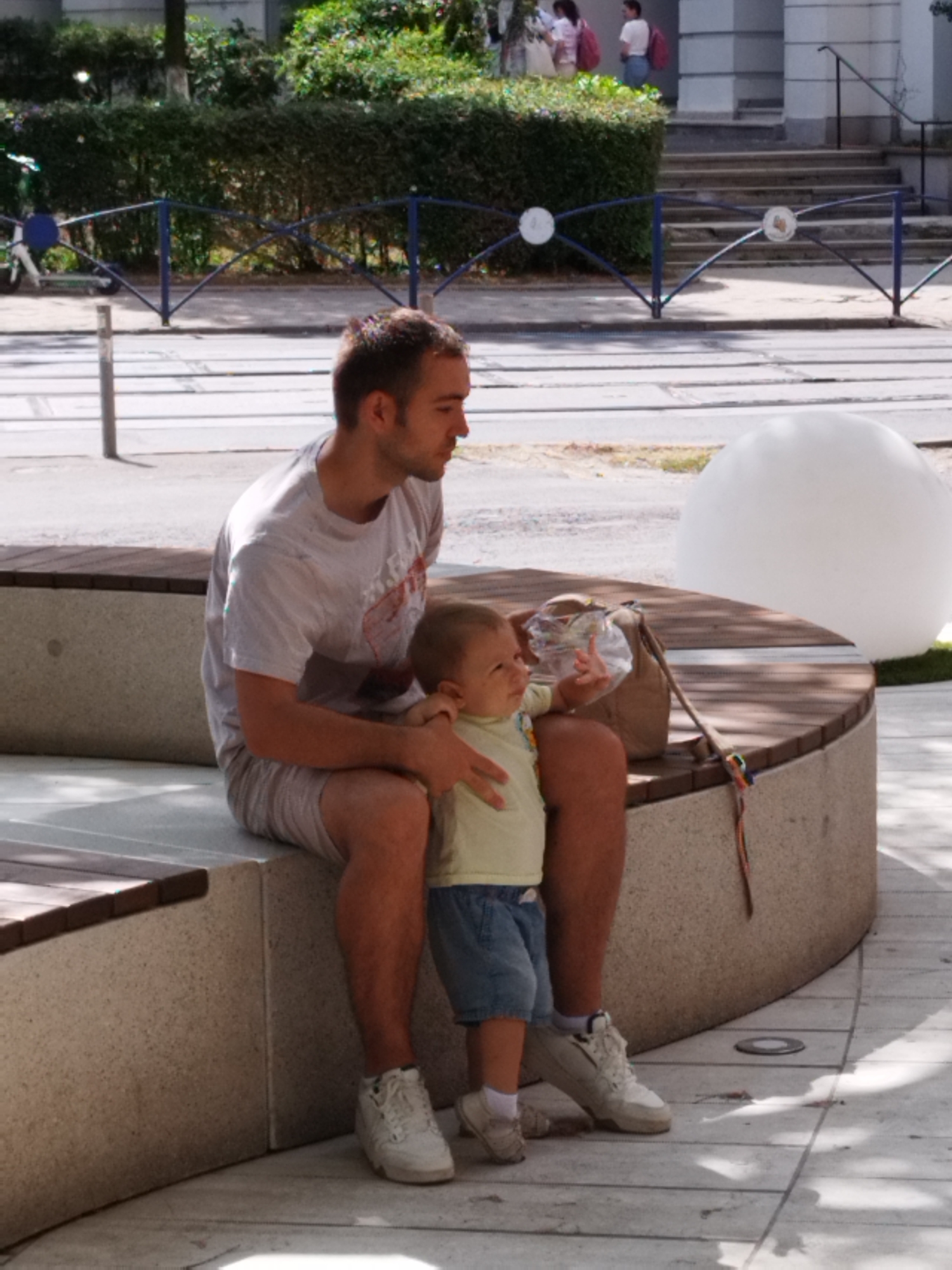 In Bucharest, Romania, a man and a baby are sitting on a curved concrete bench with wooden slats on top. The man is wearing a light-colored t-shirt and striped shorts, and white sneakers. He is holding the baby, who is wearing a light-colored shirt and blue shorts. The baby is reaching out with one hand. A brown bag and a plastic bottle are on the bench next to them. In the background, there is a large white sphere, a metal fence, trees, and a building with stairs. The ground is paved with light-colored tiles.
