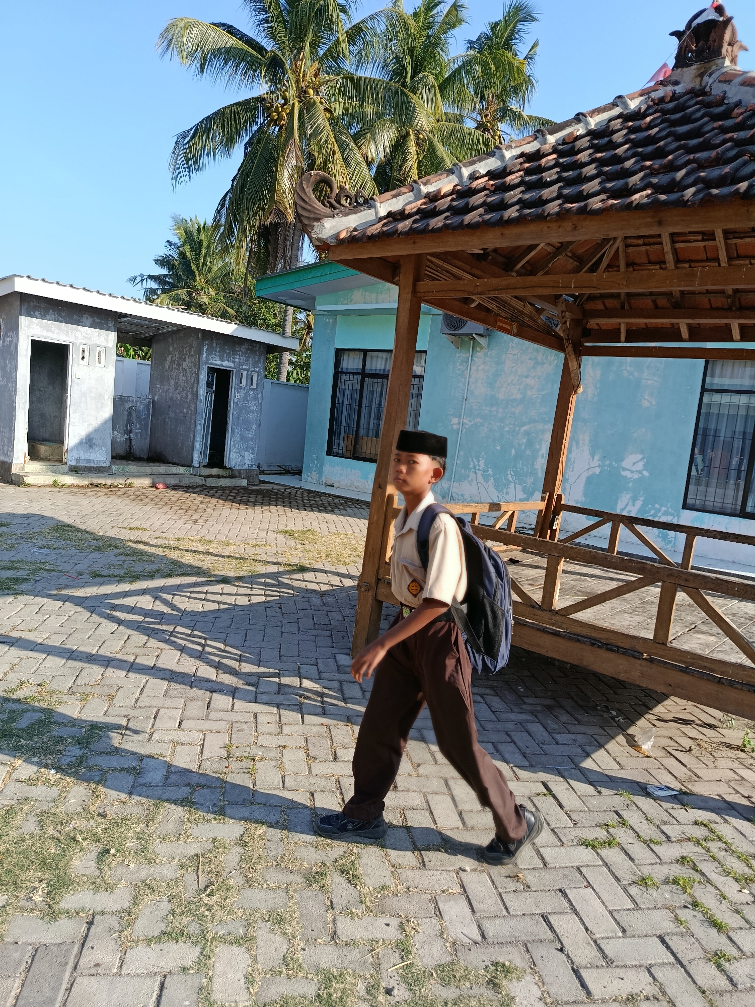 A young male student wearing a light brown uniform shirt, dark brown pants, a black peci hat, and a dark blue backpack walks across a paved courtyard in Ramban Wetan, Indonesia. The student is walking from left to right in the frame, looking slightly off-camera to the left. He is in motion, with his left leg extended forward.
In the background, there is a light blue building with dark-framed windows. To the left of the blue building are two small, gray, unpainted structures with open doorways, possibly restrooms or utility buildings. A wooden gazebo with a tiled roof, adorned with decorative elements on the roof ridge, stands to the right of the student and partially obscures the blue building.
The courtyard is paved with gray interlocking bricks, with patches of sparse green grass growing between them. Palm trees with coconuts are visible in the upper left and center of the image, indicating a tropical or subtropical climate. The scene is bathed in sunlight, casting distinct shadows from the gazebo and the student onto the paved ground. The sky above is clear and blue.