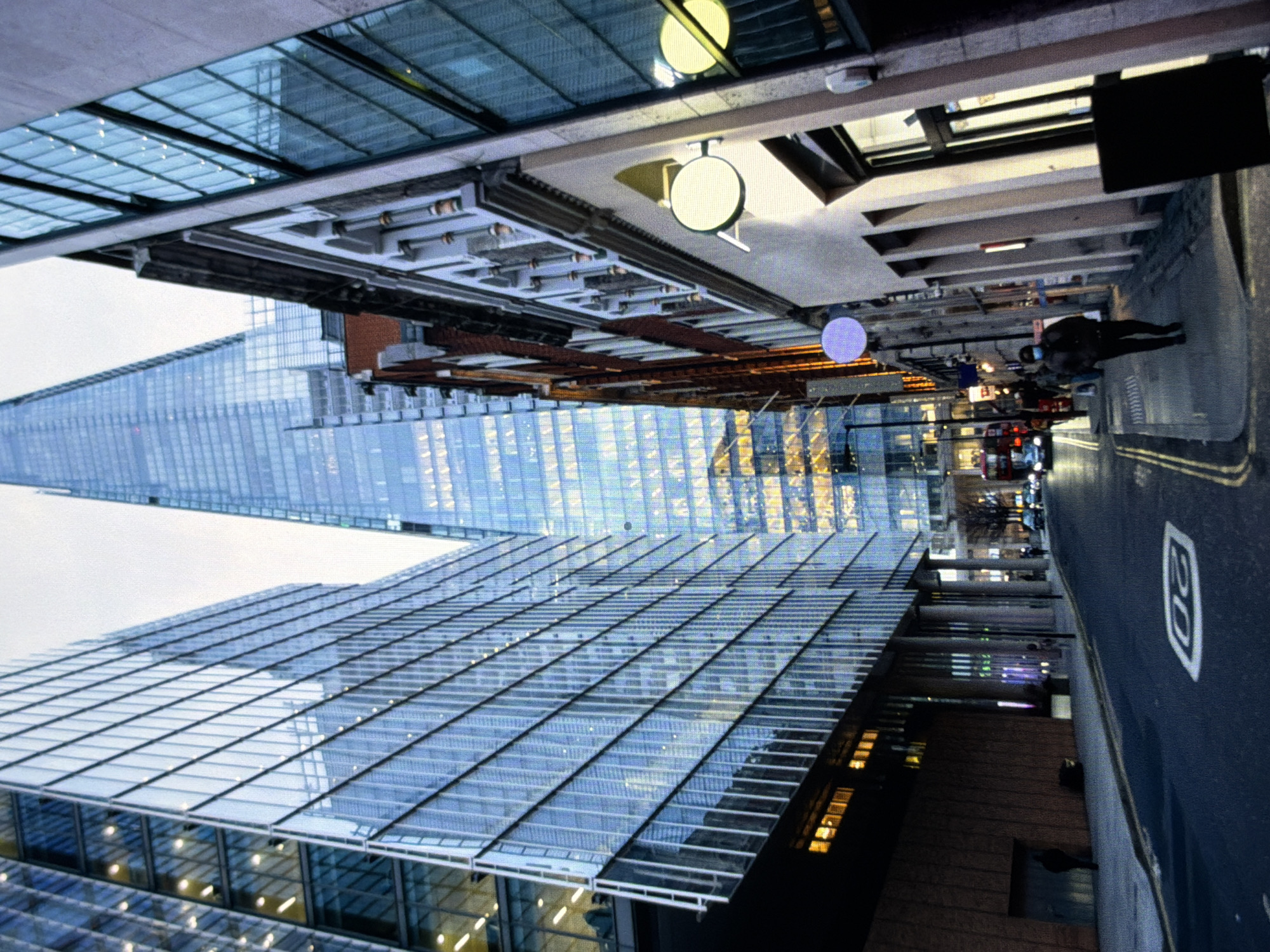 This image captures a street scene in Maidstone, United Kingdom, at dusk or during an overcast late afternoon. The view looks down a street flanked by buildings, culminating in a towering, multi-faceted glass skyscraper in the background.
On the left side of the street, modern multi-story buildings feature extensive blue-tinted glass facades that reflect the surroundings. Some internal lights are visible within these structures. The right side of the street presents a mix of architectural styles; closer buildings have lighter-colored facades with decorative elements and traditional windows, while further down, more modern glass sections are integrated. Two circular business signs, one light cream and one purple, project from a building on the right pavement.
The asphalt road in the foreground displays a white '20' speed limit marking and double yellow lines along both curbs. Pavements line both sides of the street. On the right pavement, a person wearing a dark jacket and a light blue face mask walks away from the camera. Another individual is partially visible on the left pavement further down the street, and additional distant figures can be discerned. Several cars with headlights illuminated are present on the road, and a red double-decker bus is visible further down on the left side.
The sky is overcast, and the overall lighting is dim, indicating reliance on artificial illumination from building interiors and vehicle headlights. The scene depicts a bustling urban environment characterized by a blend of contemporary glass architecture and more traditional building styles.