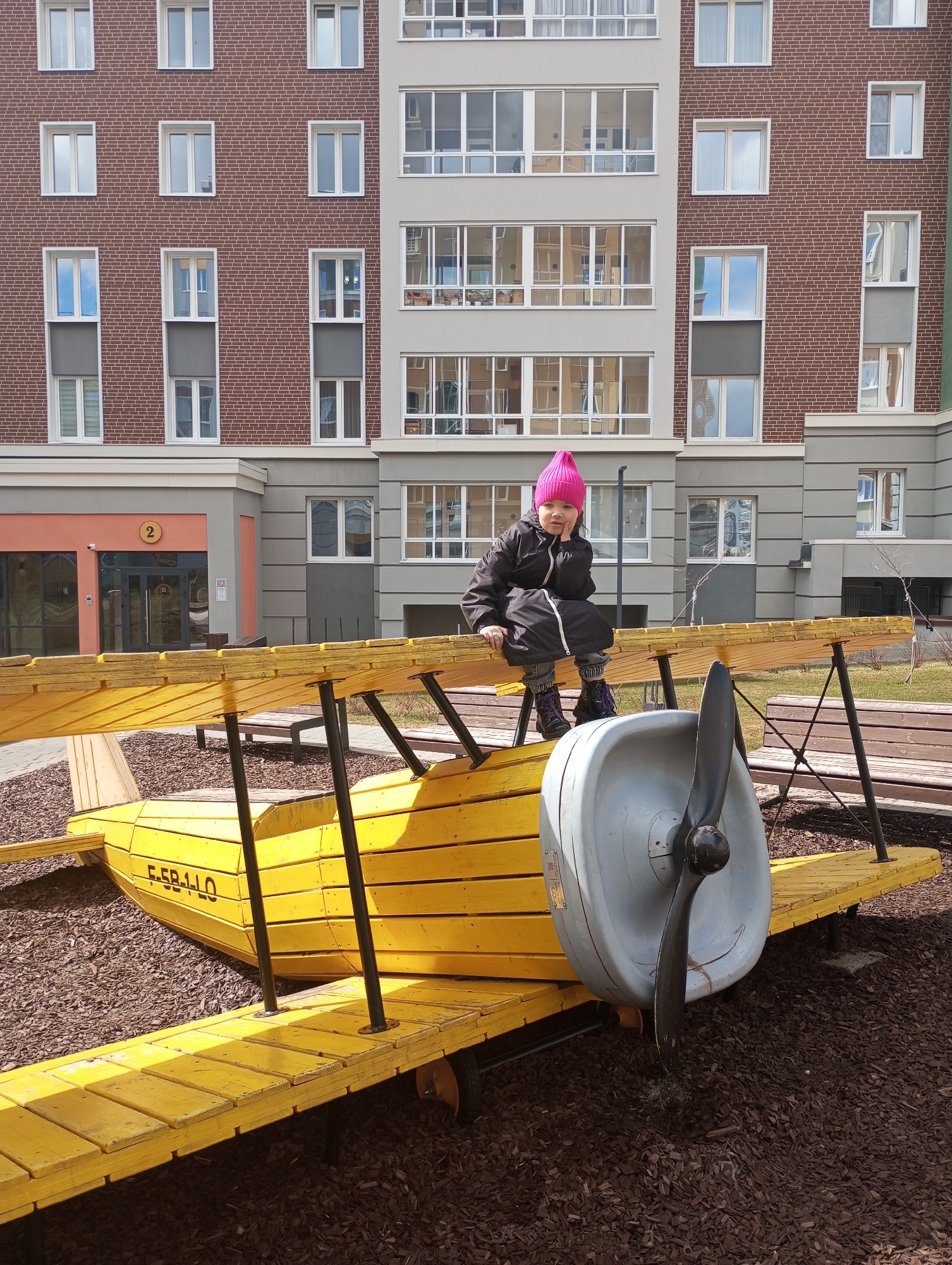 The image depicts a young girl with a pink hat sitting atop a bright yellow wooden biplane structure in a playground in Pervomaysky, Russia. She is wearing a black jacket and patterned tights. The playground surface consists of dark brown mulch.
Behind the playground equipment, a multi-story building is visible. The building features a combination of red brick and light gray facade elements, punctuated by numerous windows. The sky is reflected in some of the windows. A section of the building displays a number "2" sign adjacent to an entrance.