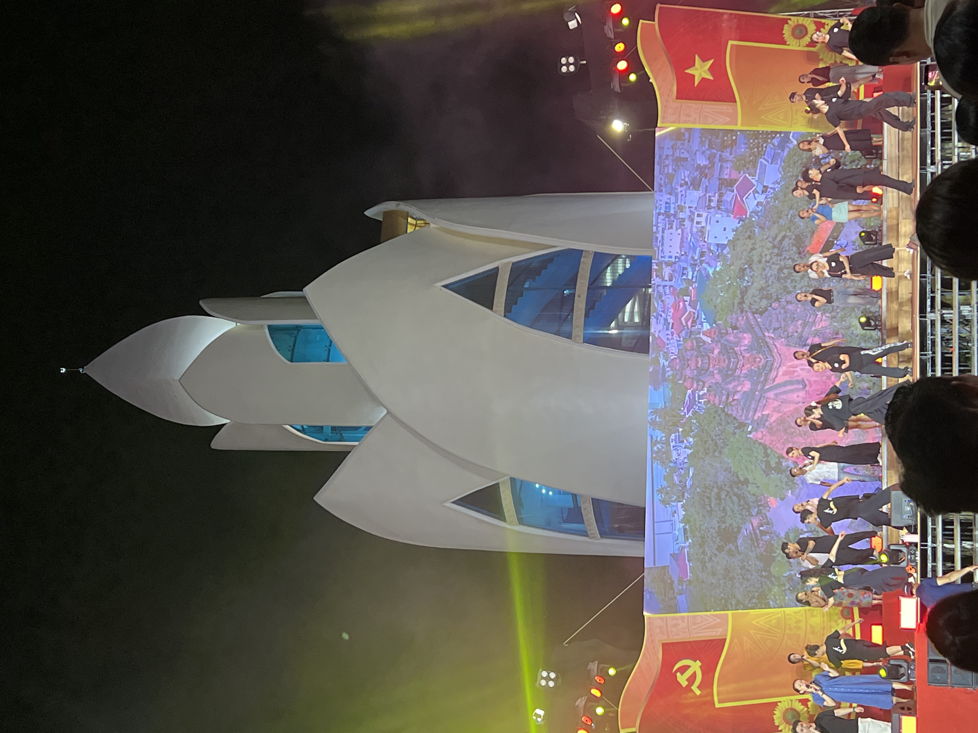 The image captures a vibrant nighttime event in Nha Trang Ward, Vietnam, featuring a distinctive modern building and an outdoor performance. On the left, a multi-layered, white building with a unique architectural design, possibly resembling a lotus flower or a futuristic structure, stands illuminated against the dark sky. This building features prominent blue-tinted windows on its various levels.
On the right, a lively stage is set for a performance. A large LED screen serves as a backdrop, displaying a colorful and detailed image of a town or village with numerous buildings and lush green landscapes. In front of the screen, a group of performers, many dressed in dark or casual clothing, are engaged in a synchronized act, likely a dance or presentation. Bright stage lights, including spotlights and colored lamps (red, yellow, white), intensely illuminate the stage area and performers, creating visible light beams and atmospheric haze.
Flanking the LED screen are two large red banners. The upper banner features the national flag of Vietnam – a yellow five-pointed star on a red field – alongside a yellow sunflower graphic. The lower banner displays a yellow hammer and sickle emblem, a symbol associated with communism, often seen in Vietnam. The heads of several audience members are visible in the immediate foreground on the far right, suggesting an engaged crowd enjoying the event. The scene evokes a festive and celebratory atmosphere during the evening hours.