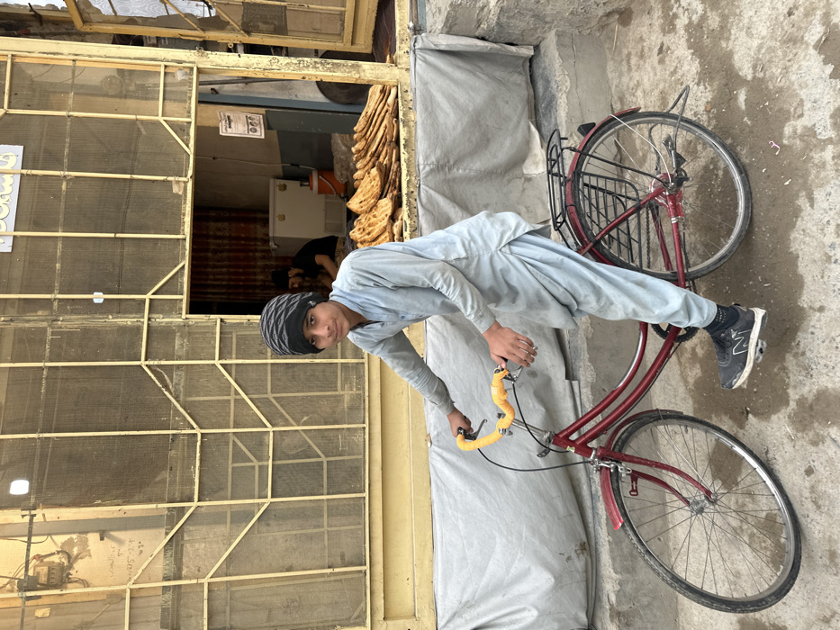 The image depicts a street scene in Quetta City Tehsil, Pakistan. In the foreground, a young male, appearing to be a teenager, is positioned on a dark red bicycle. He wears a light blue shalwar kameez, a black and grey striped beanie, and black New Balance athletic shoes. His right hand grips the right handlebar, and his left hand holds the left handlebar, which has yellow tape. His left foot rests on a pedal. He is looking directly towards the camera.
The bicycle is a step-through frame model with a rear rack and front fender. Behind the boy and the bicycle, a grey tarp or fabric is draped over a low structure. The ground in the immediate foreground is uneven concrete or dirt, with visible damp patches.
In the mid-ground, a building with a yellowish-tan facade and a security grate or screen across its front is visible. An open doorway on the right leads into a shop interior. Inside the shop, a large quantity of stacked flatbreads (likely naan) is displayed on a surface. Behind the flatbreads, a white freezer or refrigerator unit is present. A second male figure, bearded and wearing dark clothing, is partially visible inside the shop, engaged in an activity behind the counter. A white paper document with text is affixed to the wall inside the shop. Steps are visible to the right of the shop entrance.