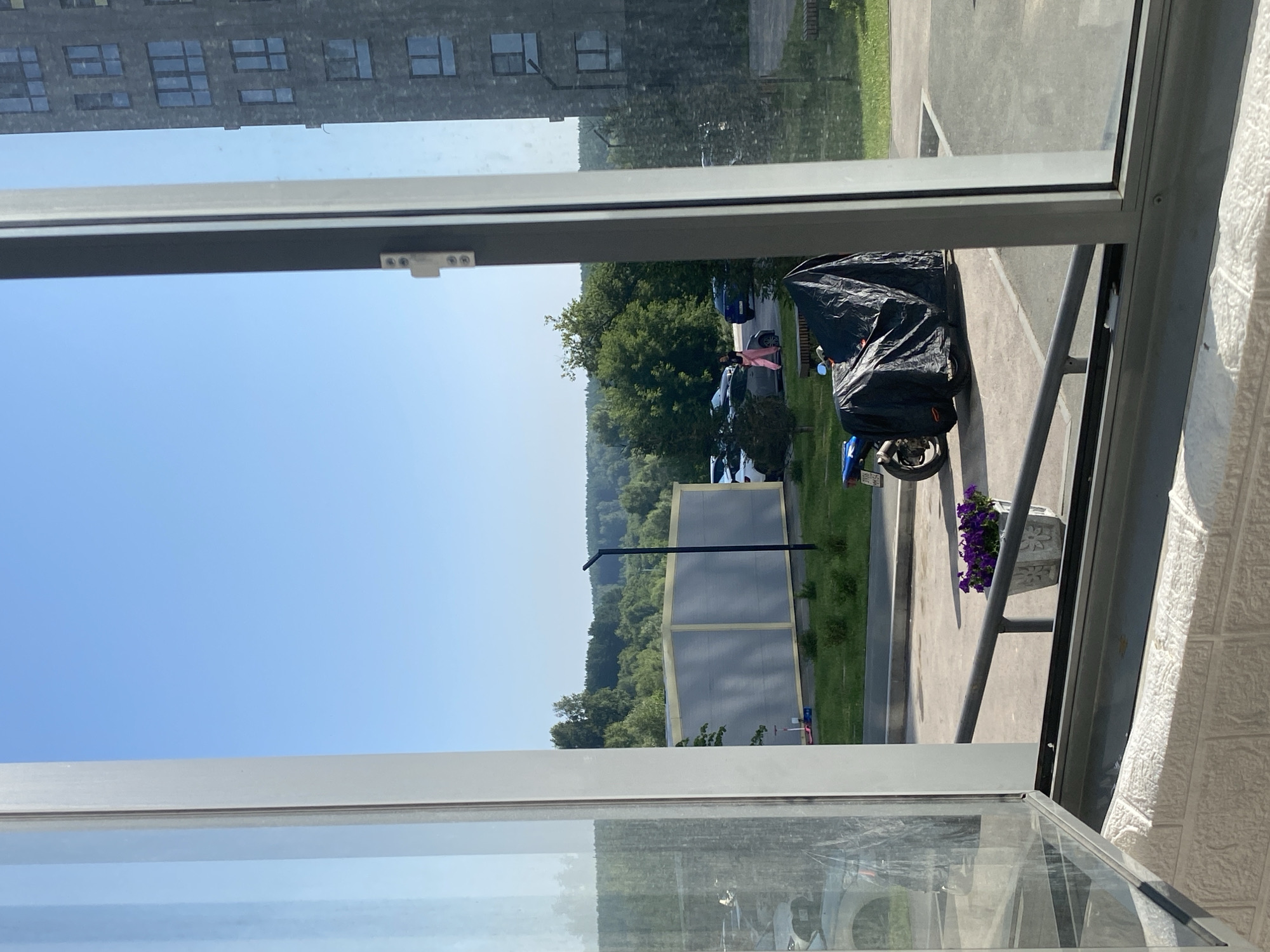 The image shows a view through a window of an apartment building in Novosibirsk, Russia. The scene depicts an outdoor area with a clear blue sky. In the foreground, an open window frame reveals a paved area with a metal railing. A motorcycle covered with a black tarp is parked on the pavement. To the right of the motorcycle is a planter with purple flowers. In the middle ground, there are trees and a light-colored building with a grey roof. Further in the distance, a forest borders the area. On the right side of the image, the facade of a dark-colored, multi-story apartment building is visible. A few cars are parked in the background, and two individuals are walking in front of some parked cars.
