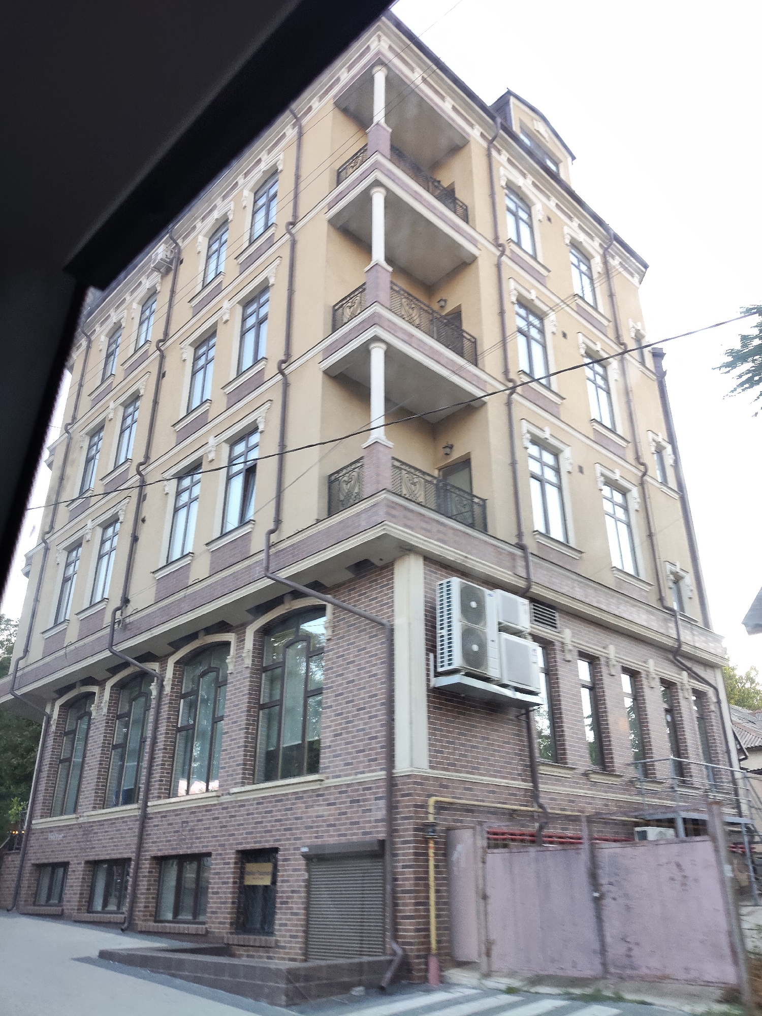 This is a photograph of a multi-story building in Chișinău, Moldova. The building is light beige with decorative white trim and a dark brown roofline. It features multiple balconies with black wrought-iron railings and white columns. The lower portion of the building is clad in dark brown brick. Several windows are visible on each floor, some of which are open. Two air conditioning units are mounted on the exterior wall on the lower floors. A metal fence and a roll-up door are visible at the ground level. Trees are visible in the background. The image appears to have been taken from a moving vehicle, as evidenced by the dark diagonal shape at the top left corner.
