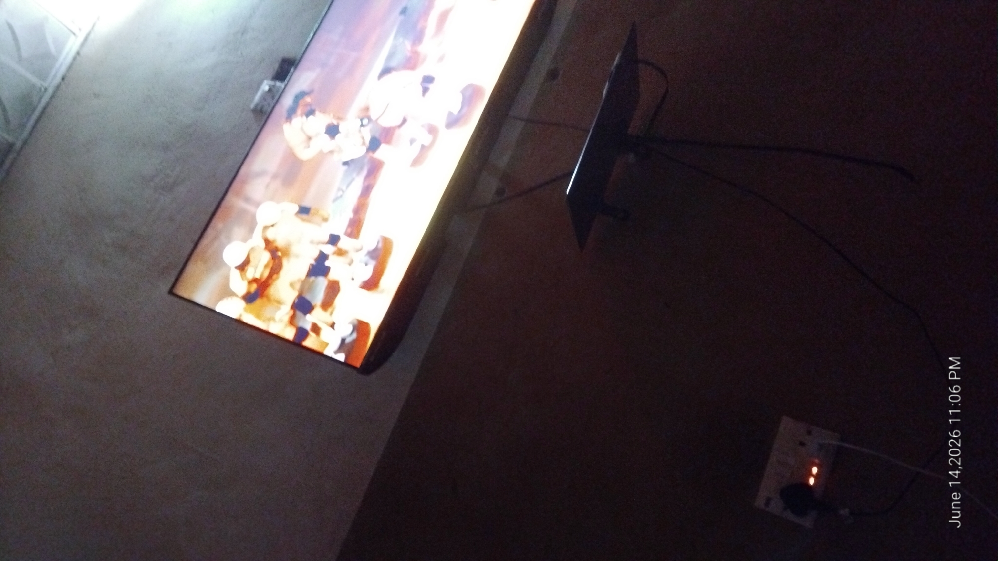 An overhead shot captures a television screen displaying a sports game with players in yellow and blue uniforms. Below the television, a black shelf is mounted on the wall. To the left of the shelf, an electrical outlet strip is visible with two illuminated orange lights and a black plug inserted. A white cord extends from the outlet strip. The image is dated "June 14, 2026 11:06 PM." The scene appears to be indoors, with a textured wall. The location is identified as Howayi, Nigeria.