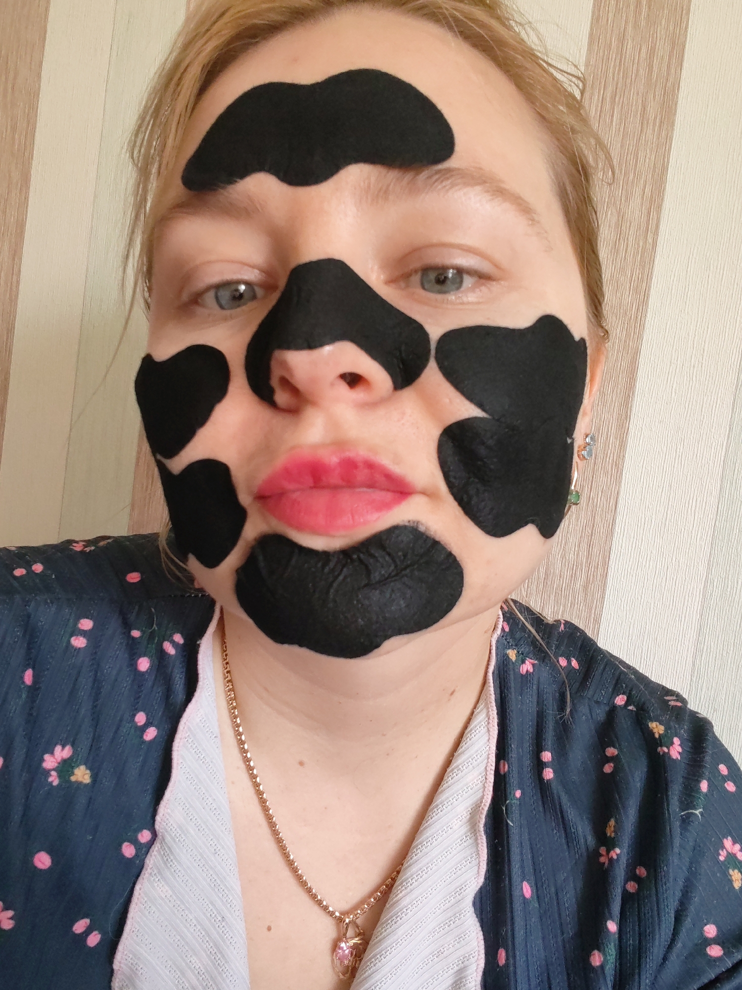 A close-up portrait shows a woman indoors in Izhevsk, Russia. She has light-colored hair, blue eyes, and pink lips. Her face is partially covered with a distinctive black face mask applied in irregular, patchy shapes, resembling a cow print or dalmatian pattern. The mask covers parts of her forehead, cheeks, nose, and chin, leaving her eyes and mouth exposed.
She is wearing a dark blue garment, possibly a robe or top, featuring a scattered pattern of small pink dots and some yellow floral accents. A light-colored trim with a ribbed texture is visible along the neckline. Around her neck, she wears a thin gold chain with a heart-shaped pendant that appears to be pink. In her right ear, a silver earring with two distinct stones, one light blue and one green, is visible. The background consists of vertically striped wallpaper in varying shades of beige and light brown. No text is visible in the image. The woman's expression is neutral.