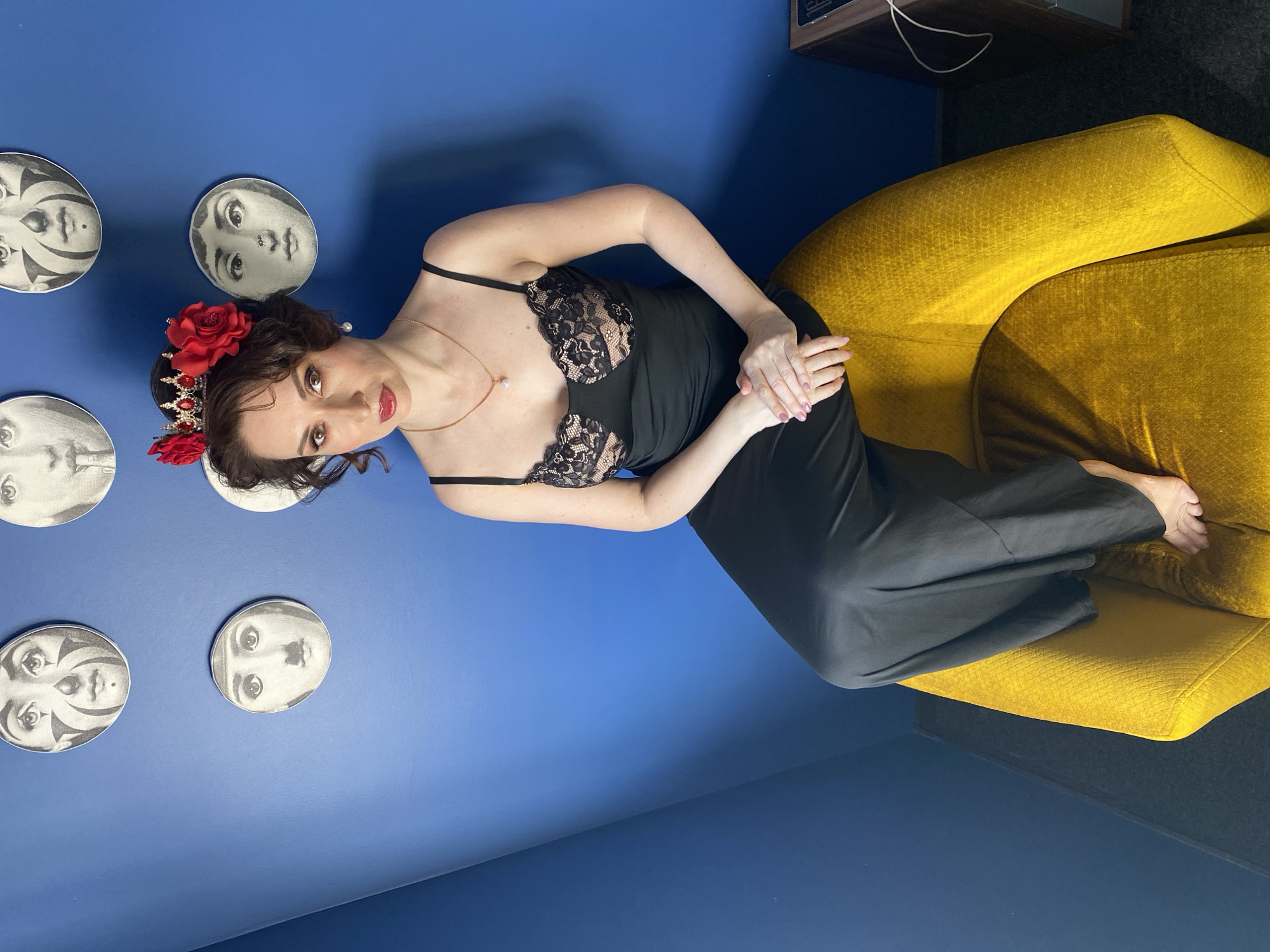 A woman wearing a black dress with lace detail sits on a yellow armchair in a room with blue walls. She has red roses and a tiara in her hair and is looking directly at the camera. The wall behind her is decorated with white face plaques. The location is Yekaterinburg, Russia. A dark cabinet is partially visible in the upper right corner of the image.