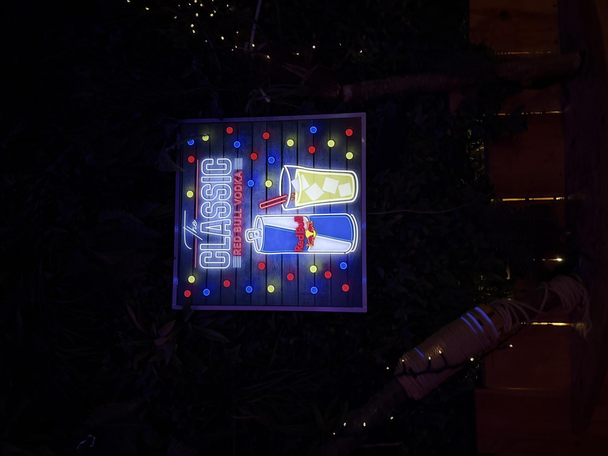 In Barcelona, Spain, a neon sign advertising "The Classic Red Bull Vodka" glows brightly against a dark, leafy background. The sign is rectangular with rounded corners and features a dark wooden plank design. In the center, the words "The CLASSIC" are written in illuminated blue neon, with "RED BULL VODKA" below in red neon. To the left of the text is an illustration of a Red Bull can in blue and white neon, and to the right is a glass with ice and a straw, rendered in yellow neon. The sign is outlined with a border of small, colorful neon dots in red, yellow, and blue. The surrounding environment is dimly lit, with foliage and lights from strings of fairy lights visible in the background. Two tree trunks wrapped in rope are present in the foreground on either side of the sign.