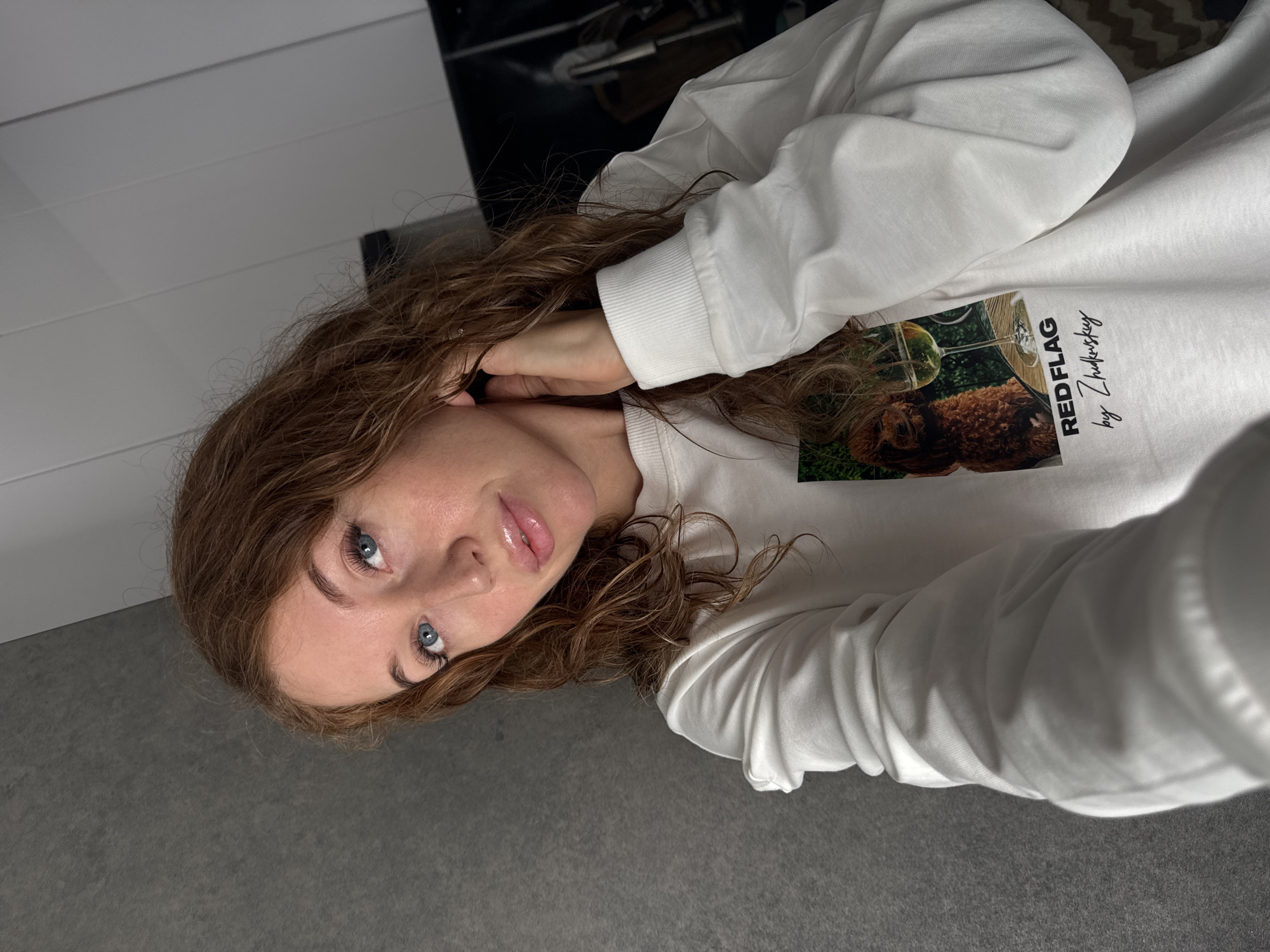 A young woman with fair skin, light blue eyes, and brown curly hair poses for a photo. She wears a white long-sleeved shirt with a graphic print of a dog and a wine glass labeled "RED FLAG by Zhukovsky." Her left hand is raised to her ear. The background includes a gray surface, possibly a floor, and a blurred black and white object in the upper background. The image was taken inside. There is no indication that this photo was taken in Chaikovsky, Russia.