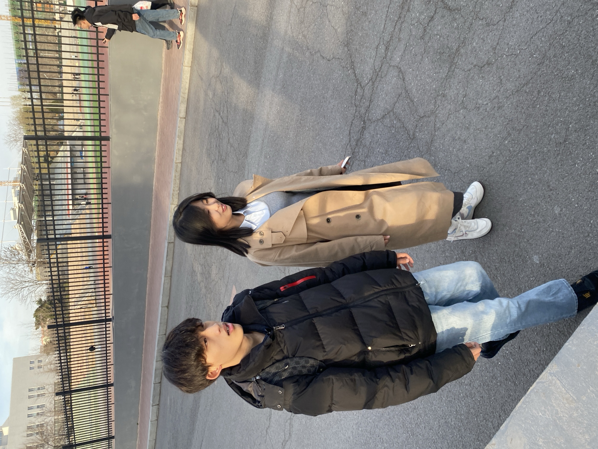 A medium-shot outdoor photograph shows a young woman and a young man standing on a paved surface near a fence. The woman is wearing a long, beige trench coat over a light-colored shirt and white sneakers. She has long, dark hair. The man is wearing a black puffy jacket and light blue jeans with black shoes. He has short, dark hair. The background includes a black fence bordering what appears to be a sports field or track. In the distance, buildings can be seen. The location is Zhongtun, China. Two blurred out people are visible beyond the fence to the upper left.