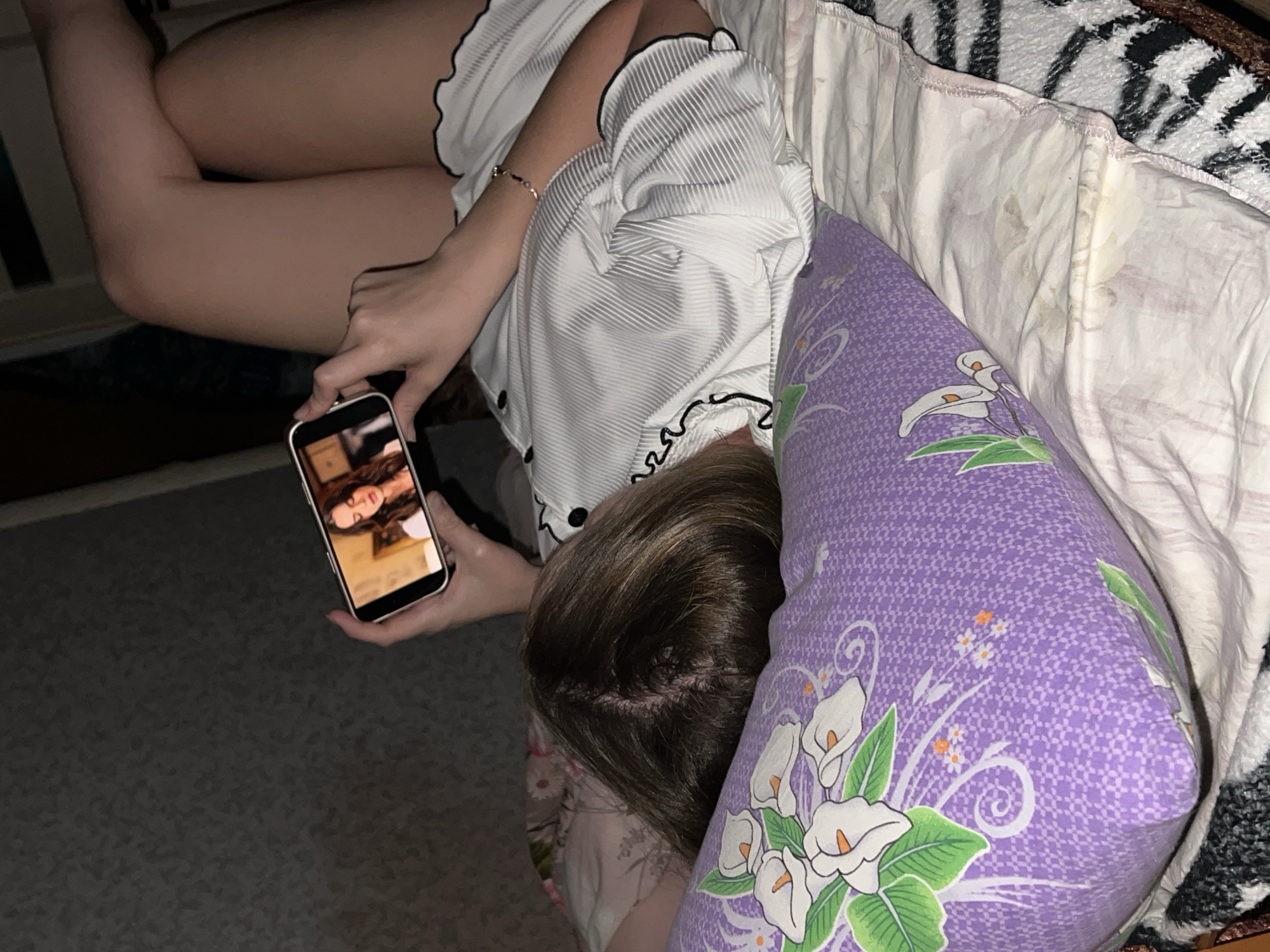 A person is reclining in a bed in an indoor environment. The person is wearing a light-colored pajama top with black trim, and their legs are visible, bent at the knees. They are holding a smartphone in their left hand, viewing an image of a face. The person's head is resting on a purple pillow with a white floral pattern. The bed has a white sheet and a blanket with a black and white zebra stripe pattern. The floor is carpeted with a neutral color. The location of the image is unknown. There is nothing in the image to suggest the location is Panteleevka, Russia.