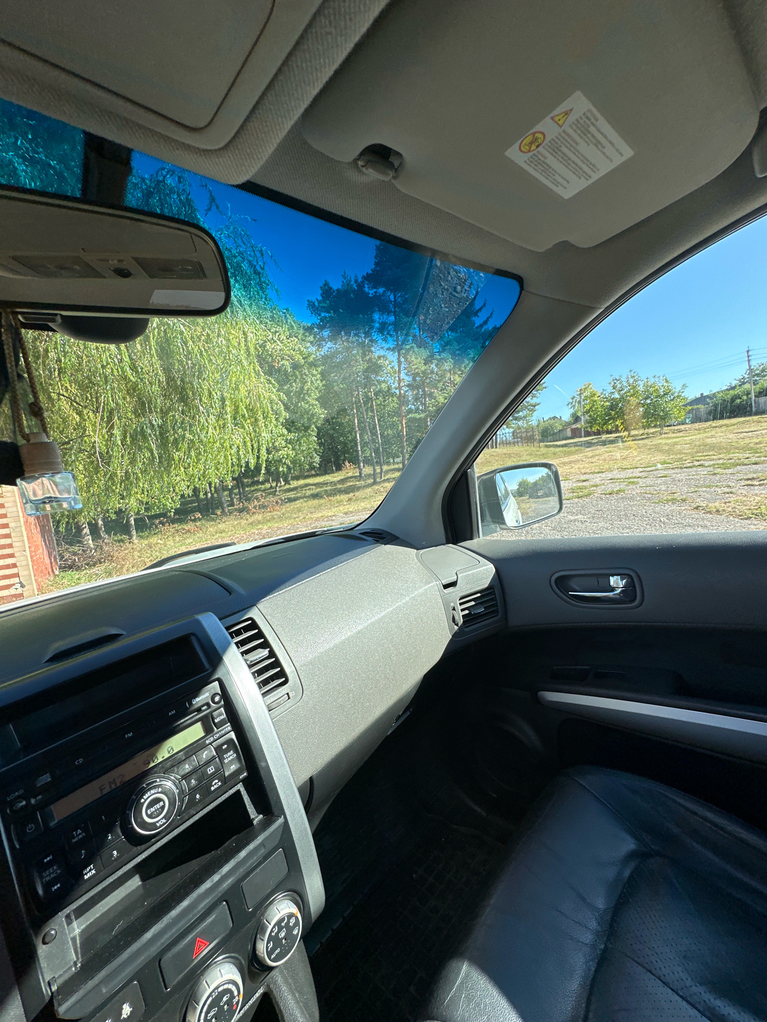 The image shows the interior of a car, looking out through the windshield and driver's side window. The car appears to be parked on a dirt or gravel lot, with trees and buildings visible in the background. The dashboard and car stereo are visible in the foreground. The sky is blue and clear. The image appears to be taken in Makiv, Ukraine, based on the metadata.