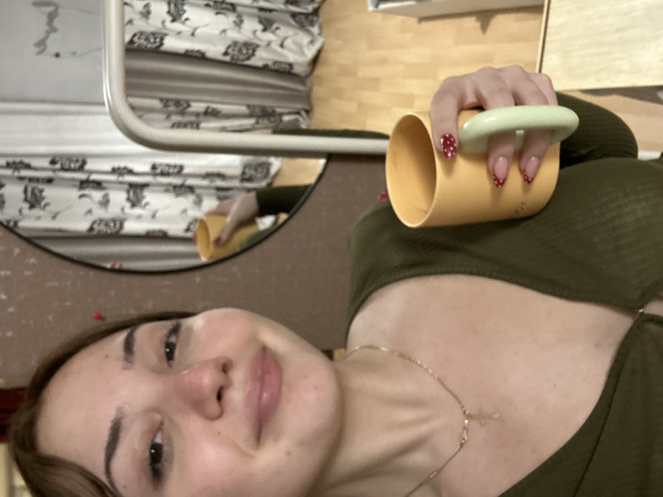 A woman with fair skin, a gold necklace, and dark hair looks directly at the camera. She holds a yellow mug with red polka dot nails. The mug has a light green handle. A round mirror is behind her with a reflection of the woman holding the mug and a bed with floral pattern bedding. The background includes a plain brown wall and a light wooden shelf.