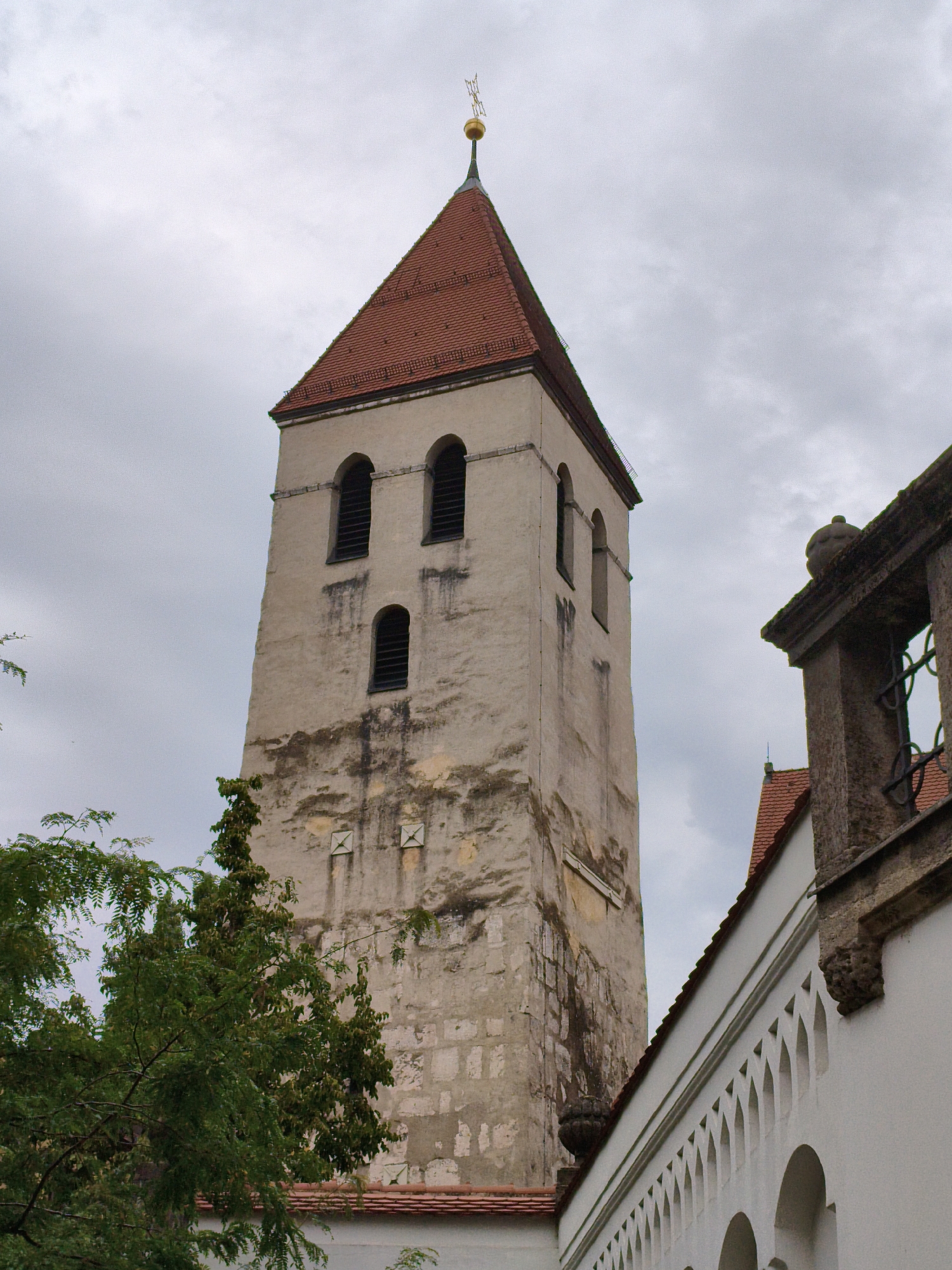 This image captures a view of a church tower in Regensburg, Germany. The tower is tall and slender, with a red tiled, steeply pitched roof topped by a golden finial. The tower's facade is made of weathered stone blocks and features several arched windows with dark louvers. Below the windows, there are two square markings on the wall. To the right of the tower, a portion of another building is visible, characterized by a white facade with a row of decorative arched openings and a stone structure with carved details on top. Lush green foliage from trees frames the lower left side of the scene. The sky is overcast with a grey, cloudy atmosphere. The overall impression is one of historical architecture under a subdued sky.