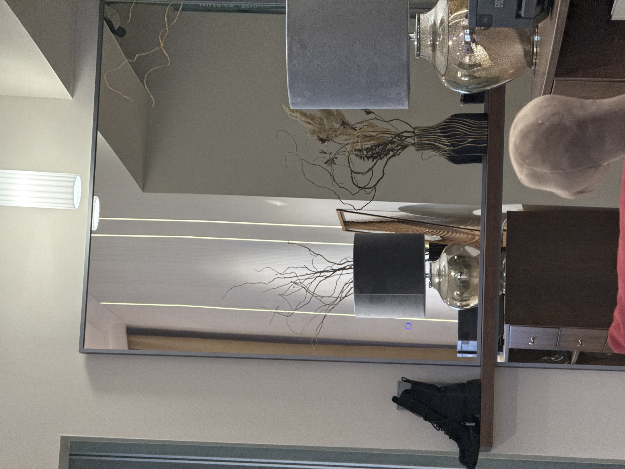 A horizontally-oriented, medium-shot photograph captures a reflected scene, seemingly indoors. The reflection is framed by a window or mirror, revealing part of an interior space. A cream-colored wall is partially visible around the edge of the frame.
In the reflection, a light fixture with a cylindrical shade is seen near the top left. Parallel lines suggest a source of indirect lighting in the room, extending horizontally. A portion of a tan curtain or panel is visible further back.
In the mirrored interior, there is a dark-colored shelf unit holding decorative items. A light gray lampshade is to the side, next to a tall glass vase. Other decor includes dried branches and dried flowers in a striped vase. A partially visible item that looks like a wig head. Below, another similar decorative arrangement is visible with another lampshade.
Near the bottom, a dark-colored boot is visible. This suggests the presence of a hallway or other entryway nearby.
The location and city of Nizhnevartovsk, Russia, cannot be determined from this information.