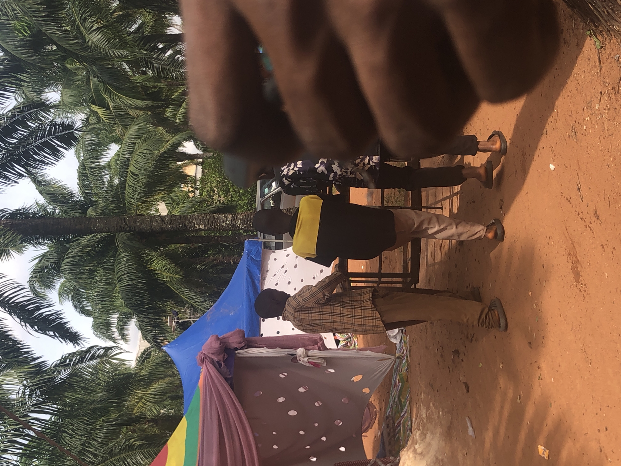 The scene is set in Nteje, Nigeria, with palm trees visible in the background. There are three individuals present, though one is heavily obscured by what appears to be a hand held close to the camera. The other two individuals stand near a makeshift structure covered with colorful fabric and a dotted white sheet. One person wears a plaid shirt and khaki pants, while the other wears a black shirt with a yellow stripe and light-colored pants. The ground is reddish-brown dirt.