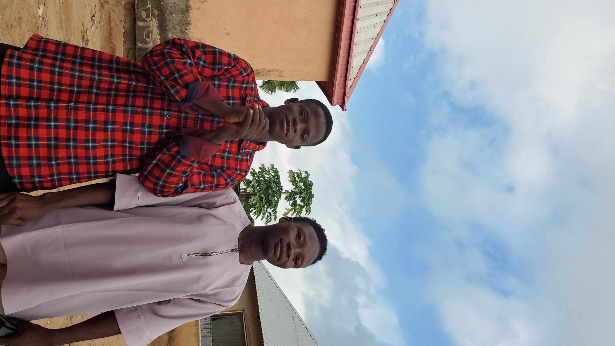 Two young men stand outside, posing for a photograph. The man on the left wears a red and black plaid shirt with the sleeves rolled up and his hands clasped in front of him. The man on the right wears a light purple, loose-fitting garment with a zipper closure at the neck. They are positioned in front of a simple, peach-colored building with a red-trimmed roof. To their right, another building with a grey, corrugated roof is visible. A tree with broad green leaves stands between the two men. The ground is dirt, and the sky above is a mix of blue and white clouds. The scene appears to be in a residential area of Jalingo, Nigeria.
