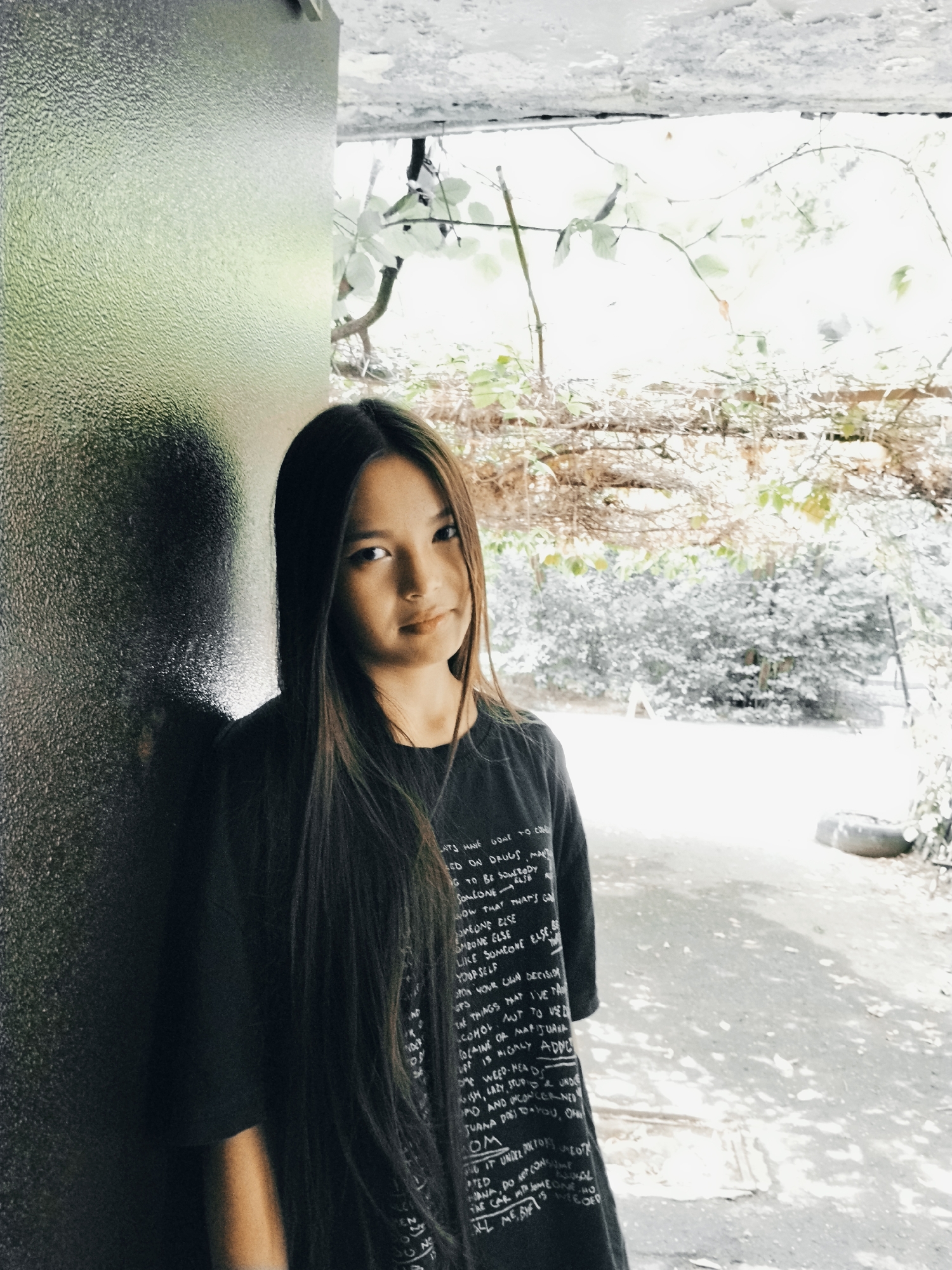 A young woman with long, dark hair stands in an outdoor setting, leaning against a textured, dark gray surface on the left. She is wearing a black t-shirt with white text printed on it, which is partially visible. Her gaze is directed slightly to the right of the camera. The background consists of foliage, branches, and a concrete ground, suggesting an urban environment. The lighting indicates it is daytime, with dappled sunlight creating shadows on the ground and the woman's clothing. The overall scene appears to be an alleyway or a secluded outdoor area within Bishkek City, Kyrgyzstan, possibly near a building's exterior wall.