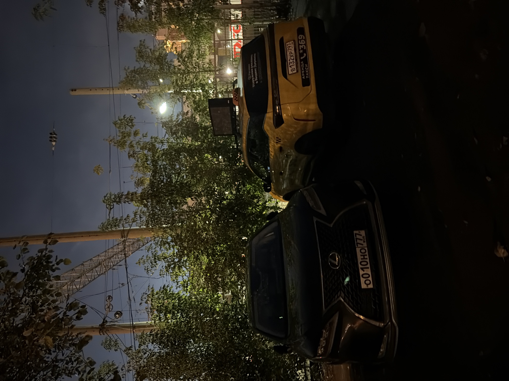 An exterior, medium shot captures two parked cars at night, with a dark sky above. On the left is a dark-colored Lexus SUV, its front grille and license plate clearly visible. The license plate reads "0010HO 777". To its right is a yellow taxi, also an SUV, featuring the text "B132XP 797" and "*369" on its rear license plate. The taxi also has a "TAKSI" label on its side. The vehicles are parked near a dense growth of green trees and foliage. In the background, a dimly lit building and streetlights are visible. Power lines and poles traverse the scene, and a large metallic structure, possibly a pedestrian bridge or scaffolding, is suspended above the cars. The overall lighting suggests an urban environment in Moscow, Russia.